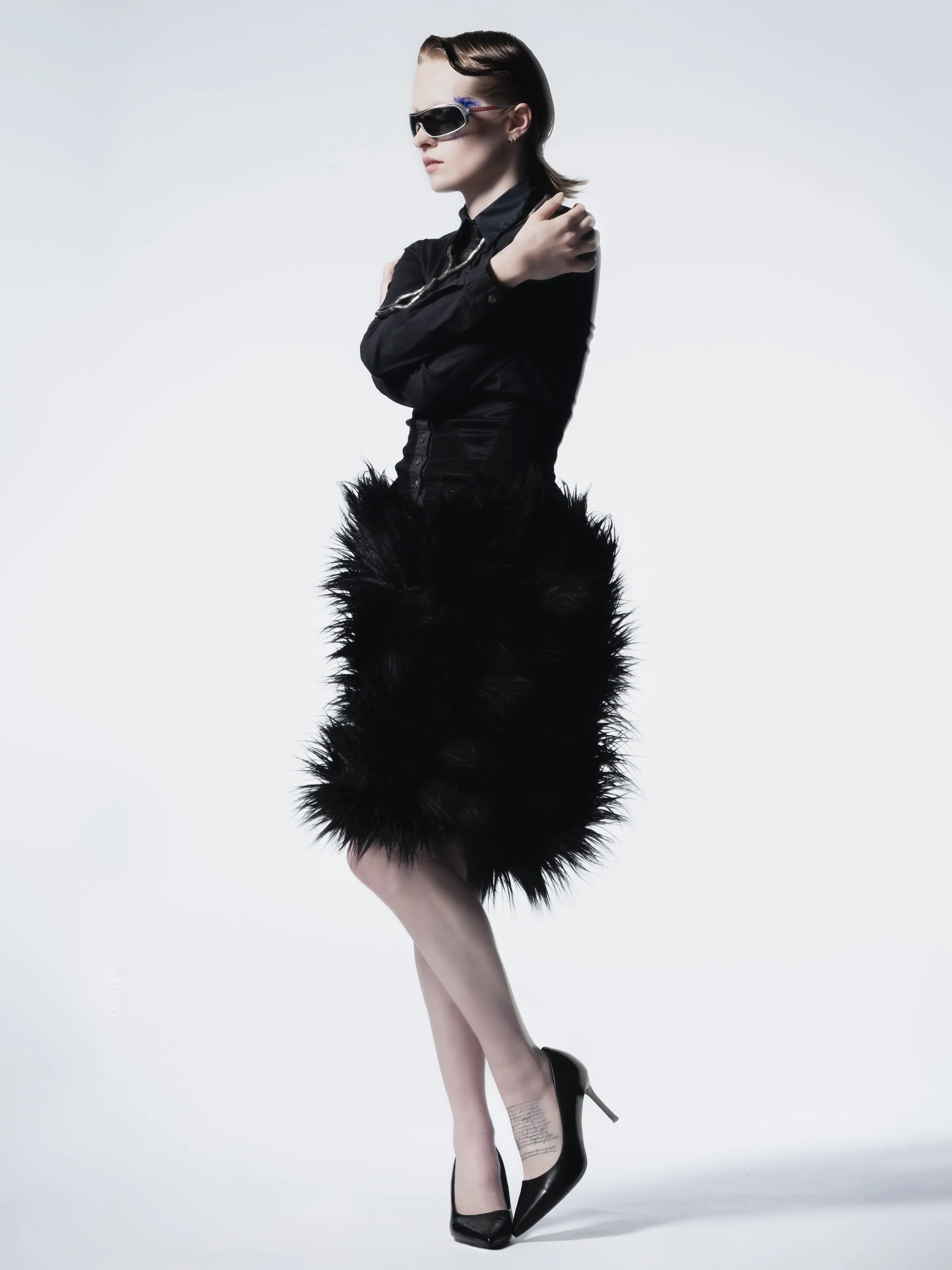 Femme portant des lunettes de soleil noires, une robe noire en haut, une jupe en plumes noires, chaussures à talons noirs, posant contre un fond blanc.