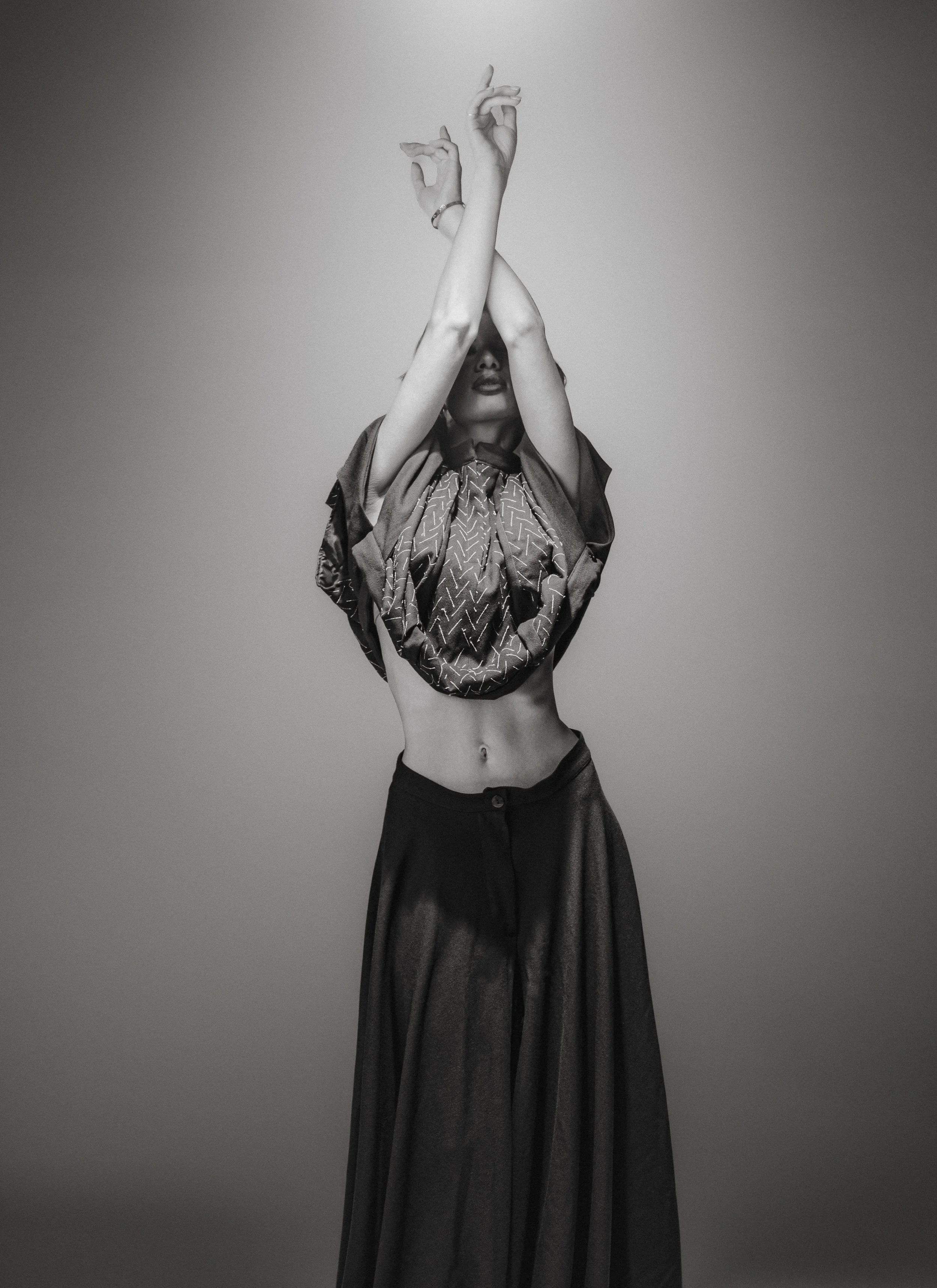Une femme en noir et blanc avec des bras croisés au-dessus de la tête, portant un haut ample et une longue jupe, se tenant contre un fond uni.