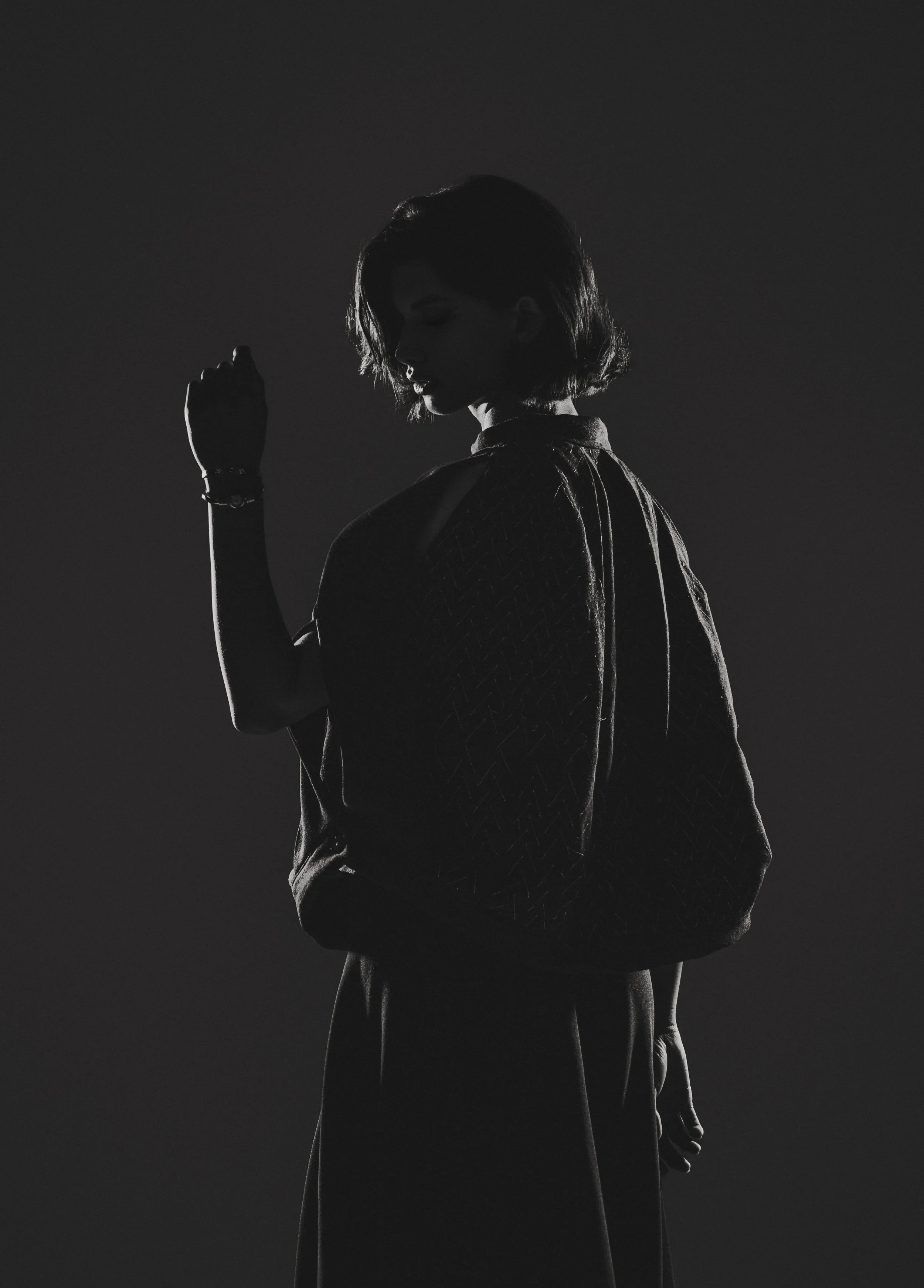 Silhouette d'une femme en noir et blanc, vue de dos, avec les bras pliés, portant un sac à dos et une montre, dans un environnement sombre.
