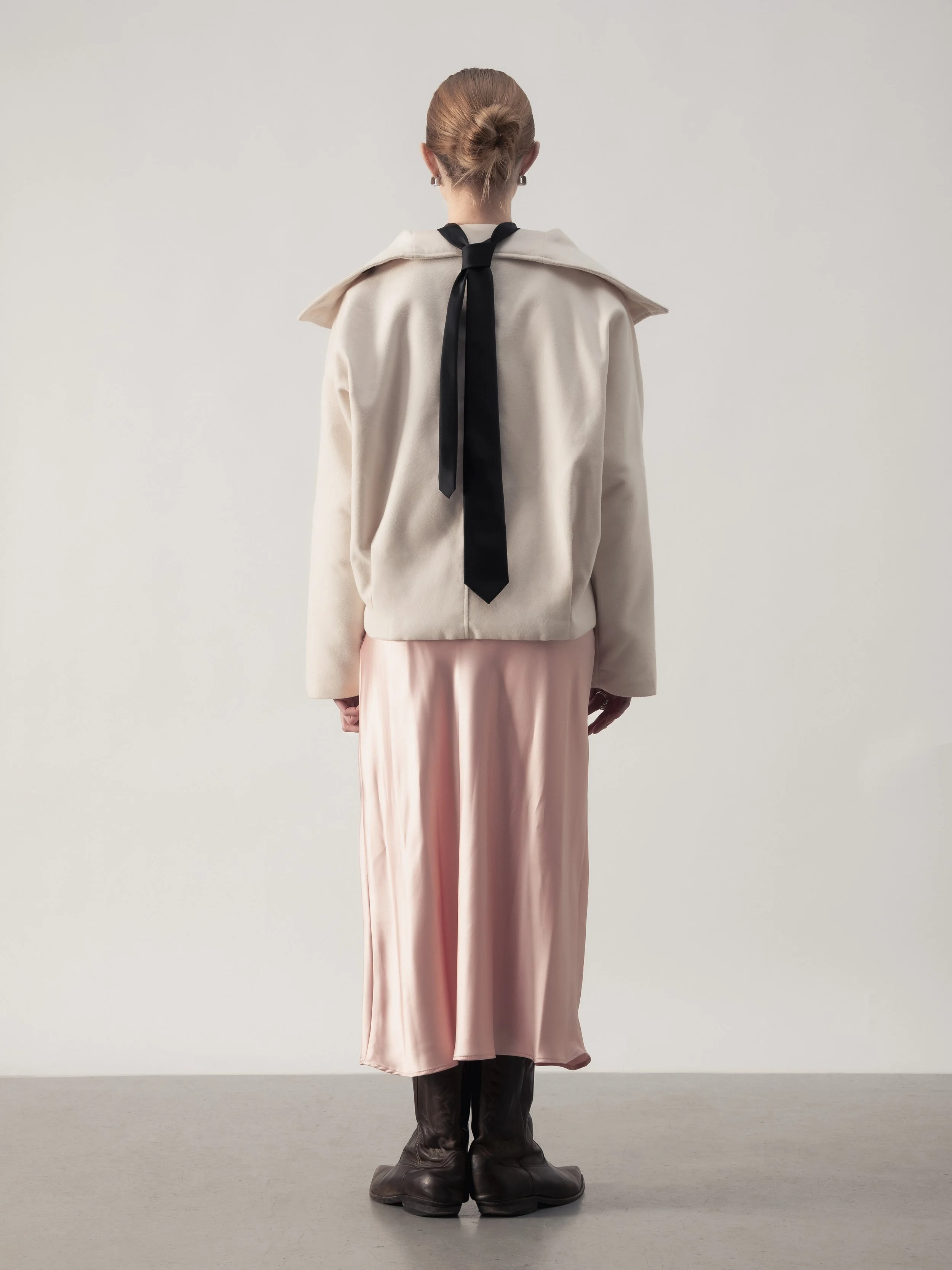 Femme vue de dos, portant un manteau beige, une jupe longue en satin rose et des bottes en cuir marron.