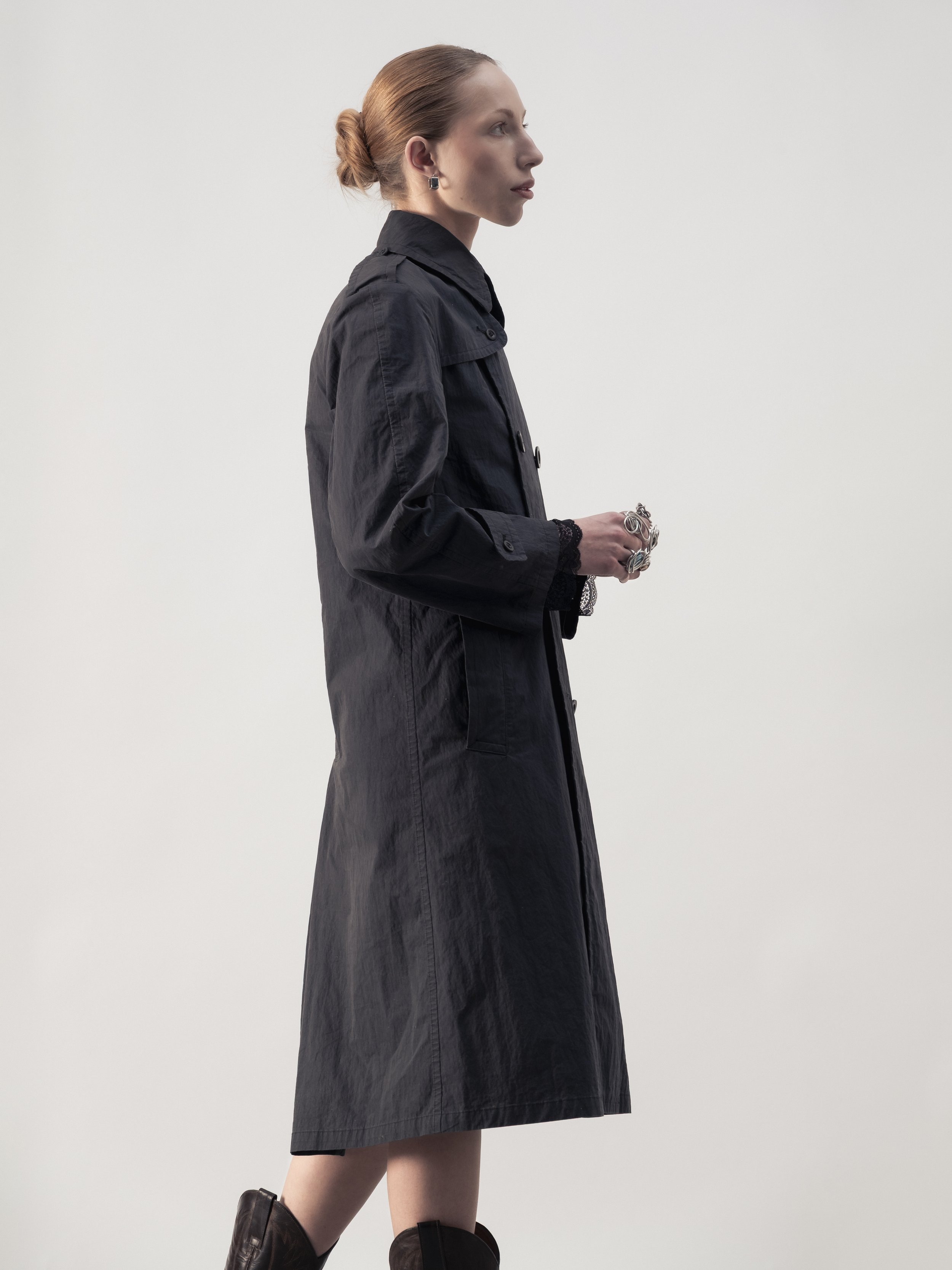 Femme en profil portant un manteau long noir, accessoires en argent, et bottes marron, avec un fond blanc.