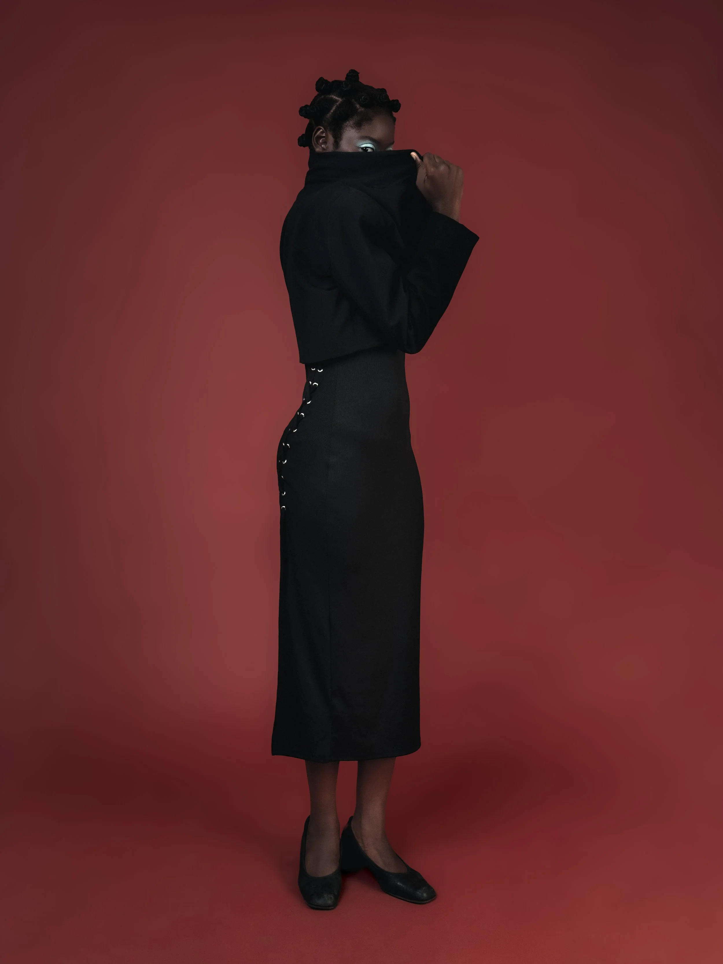 Femme portant un haut noir et une jupe longue, cachant partiellement son visage, avec un fond rouge.