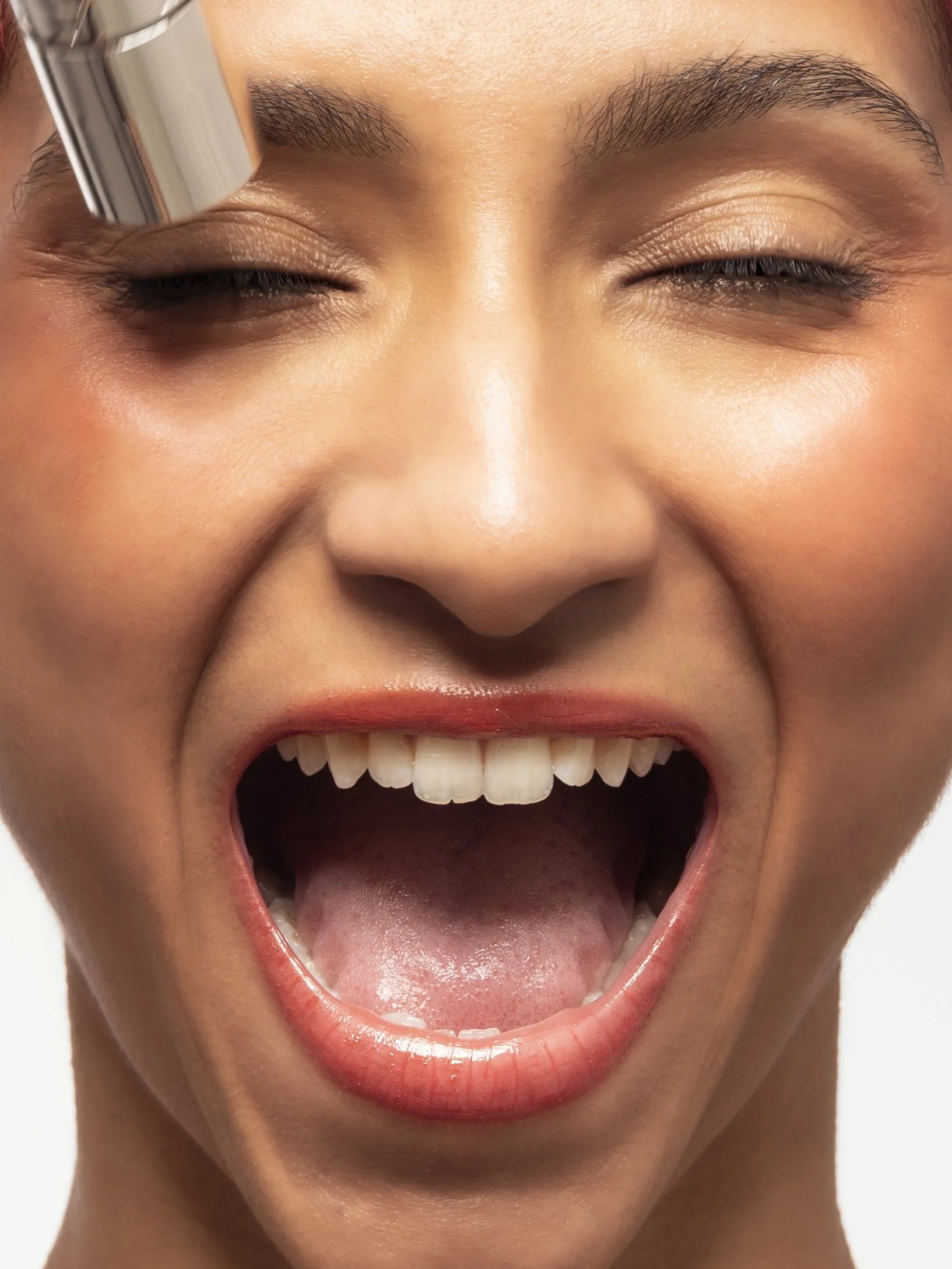 Une femme souriante avec les yeux fermés, montrant ses dents, en train d'utiliser un instrument de beauté sur son œil.