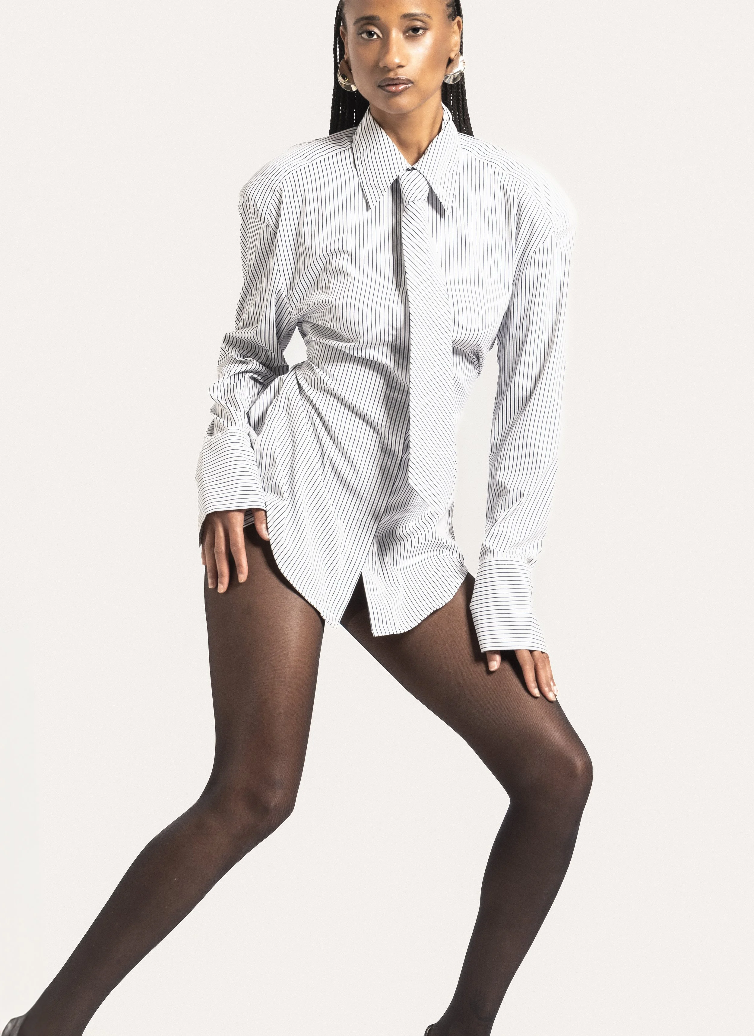 Une femme porte une chemise blanche à fines rayures noires, une cravate assortie, et des collants noirs, posant devant un fond blanc.