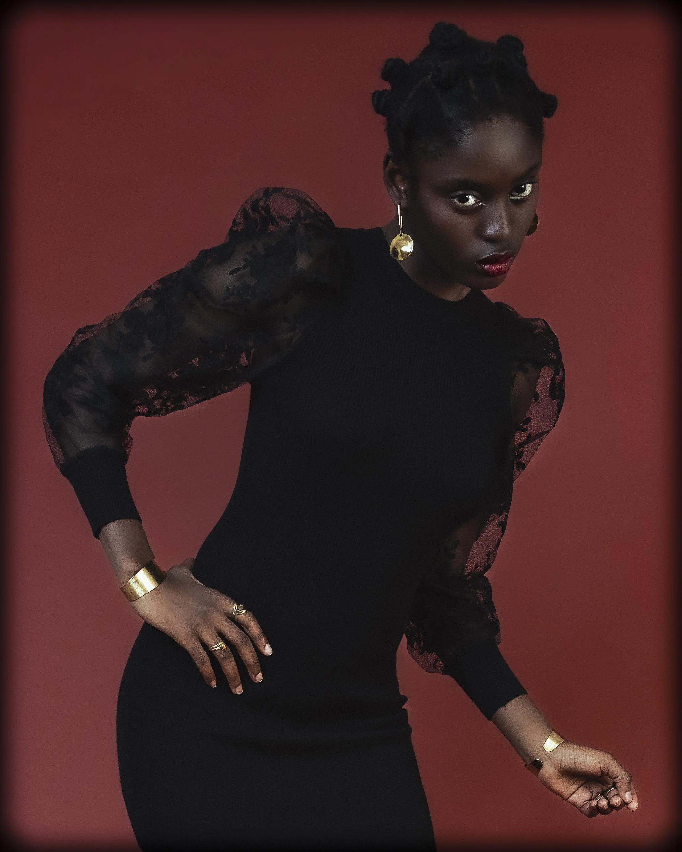 Une femme noire portant une robe noire avec des manches en dentelle transparente et des bijoux dorés, posant contre un fond rouge.