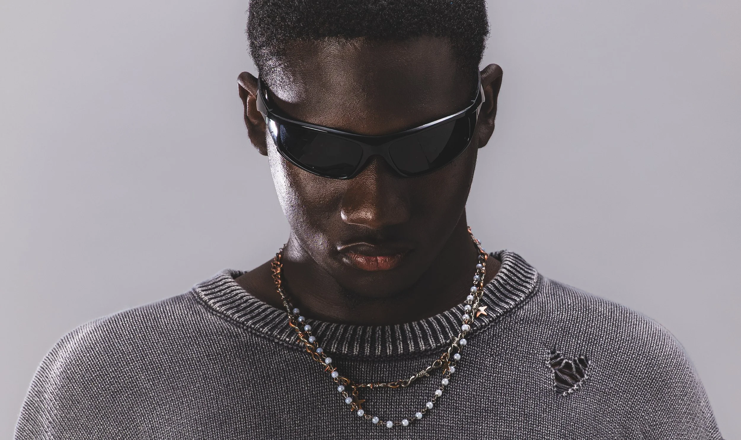 Jeune homme noir portant des lunettes de soleil noires, un pull gris avec un trou en forme de cœur, et deux colliers, contre un fond gris clair.