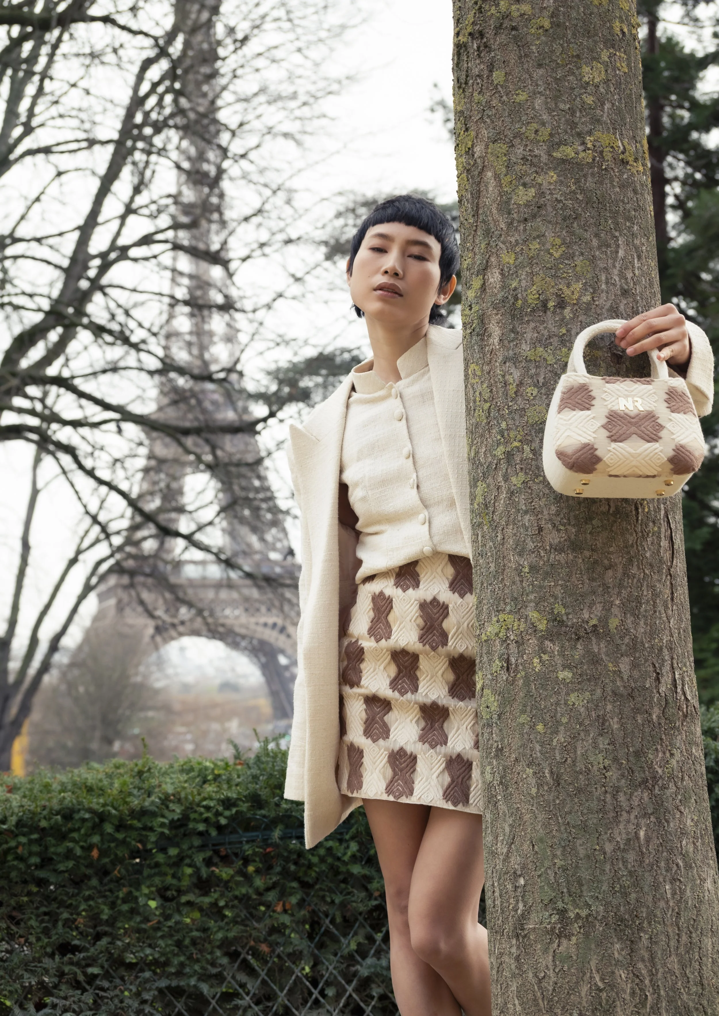 Une femme posant derrière un arbre avec la tour Eiffel en arrière-plan, portant une tenue beige avec un sac à main beige et marron.