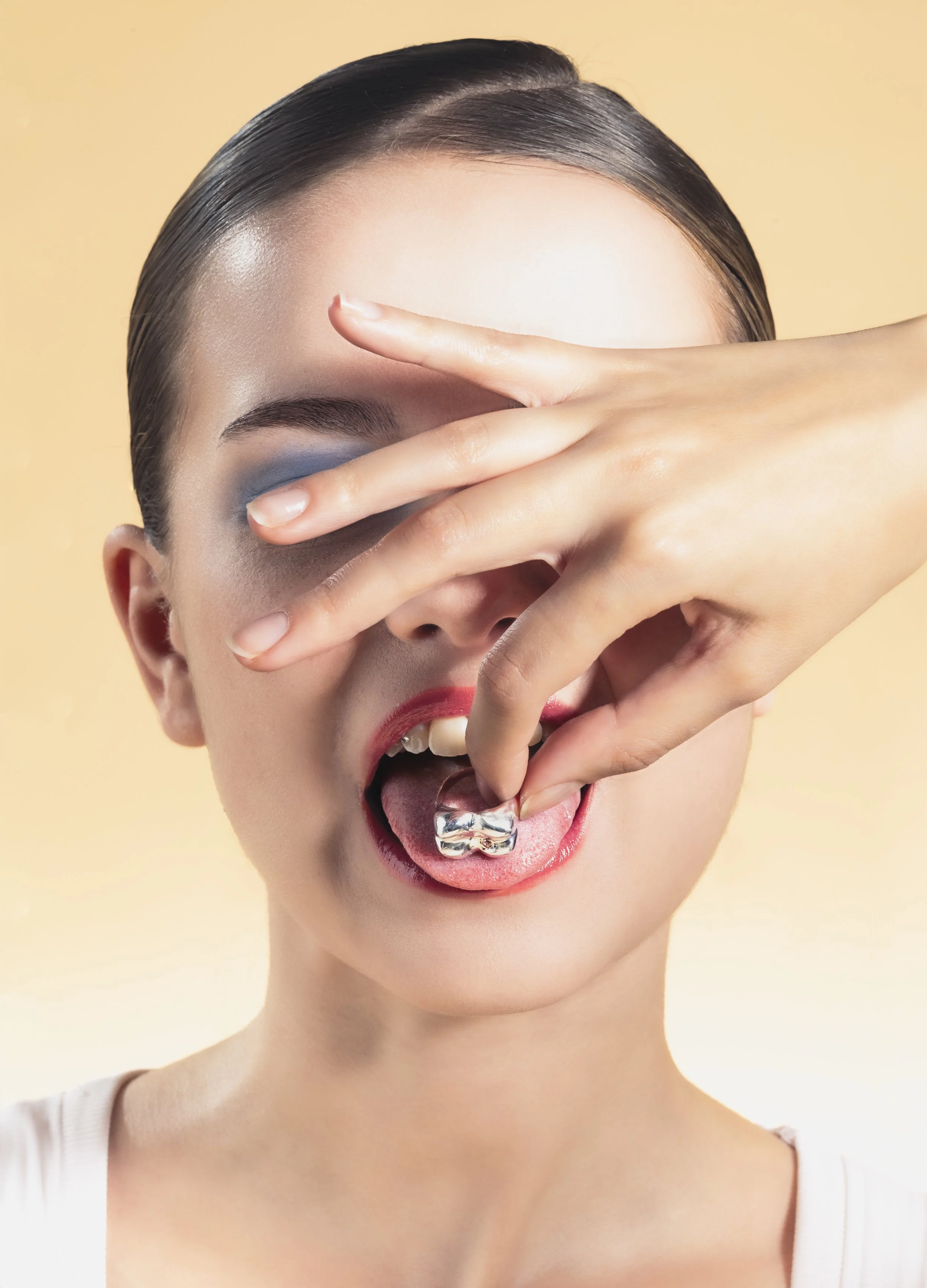 Jeune femme avec maquillage coloré, portant un lingot d'argent sur la langue, faisant un geste artistique avec ses doigts devant son visage contre un fond beige.