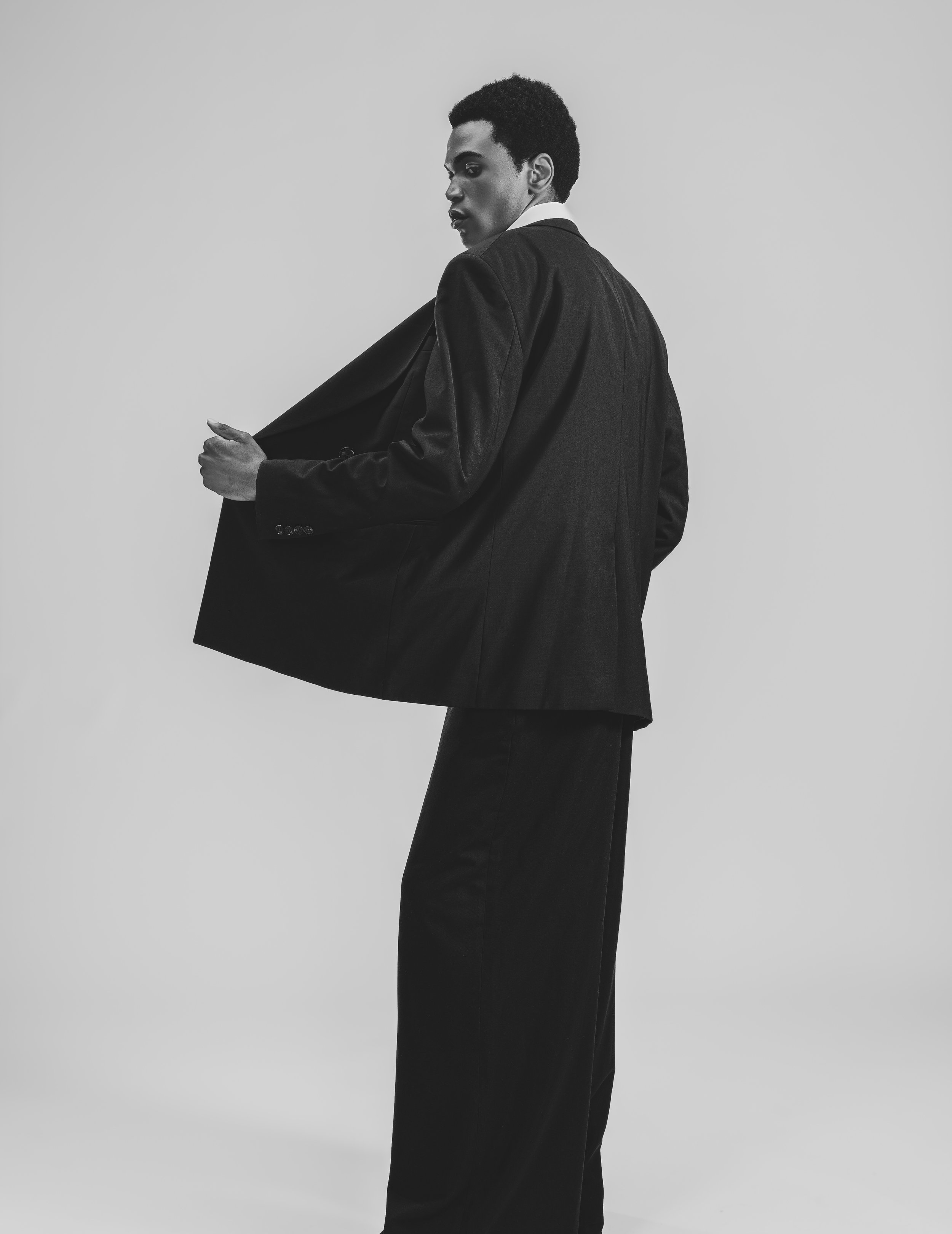 Un homme habillé en costume noir avec une chemise blanche, qui tient la poche de son blazer, regardant vers la gauche, en noir et blanc.