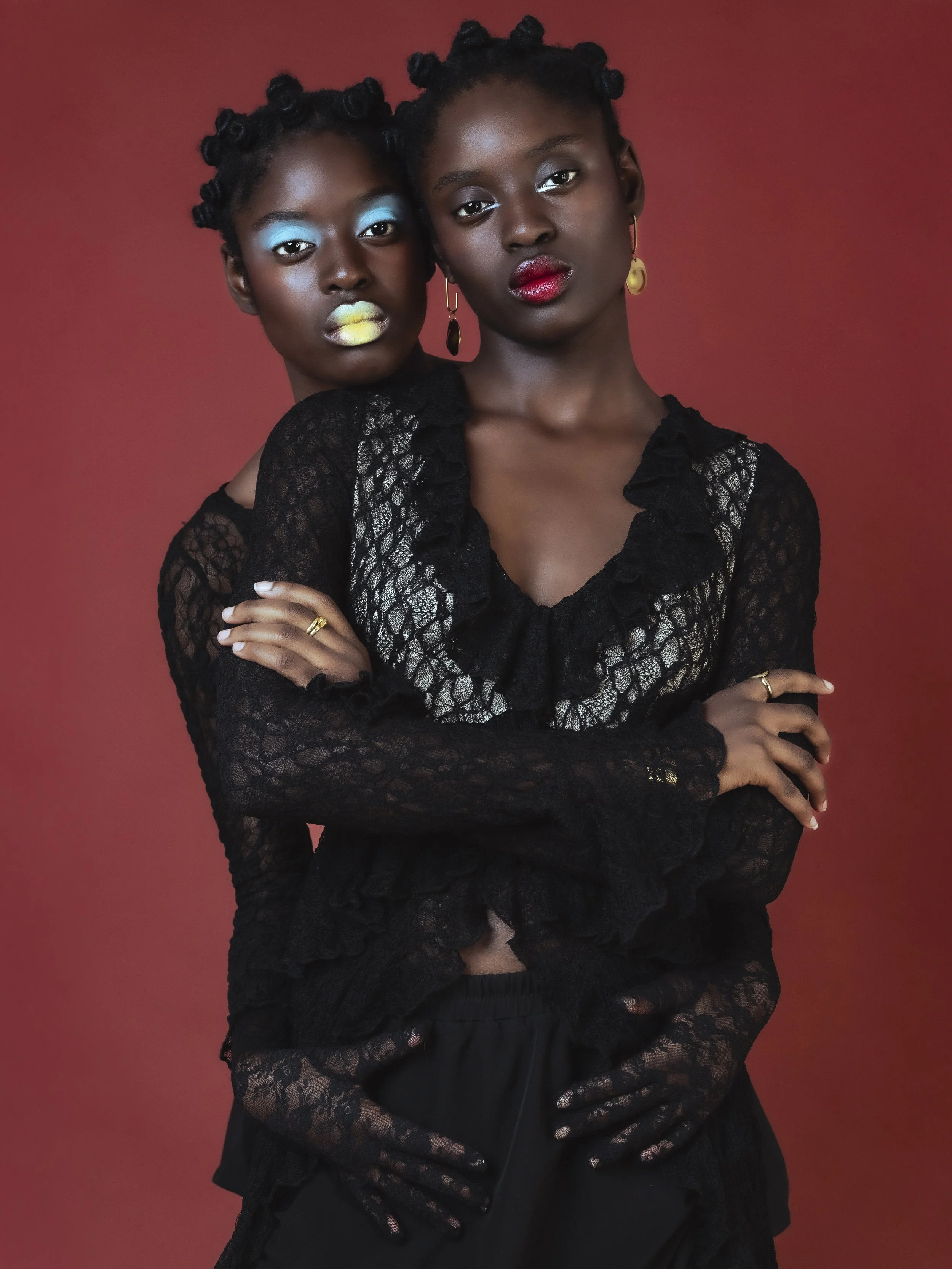 Deux femmes africaines posant contre un fond rouge, portant des vêtements en dentelle noire, maquillage coloré et boucles d'oreilles. L'une a des lèvres jaunes et bleues, l'autre a du rouge à lèvres rouge.