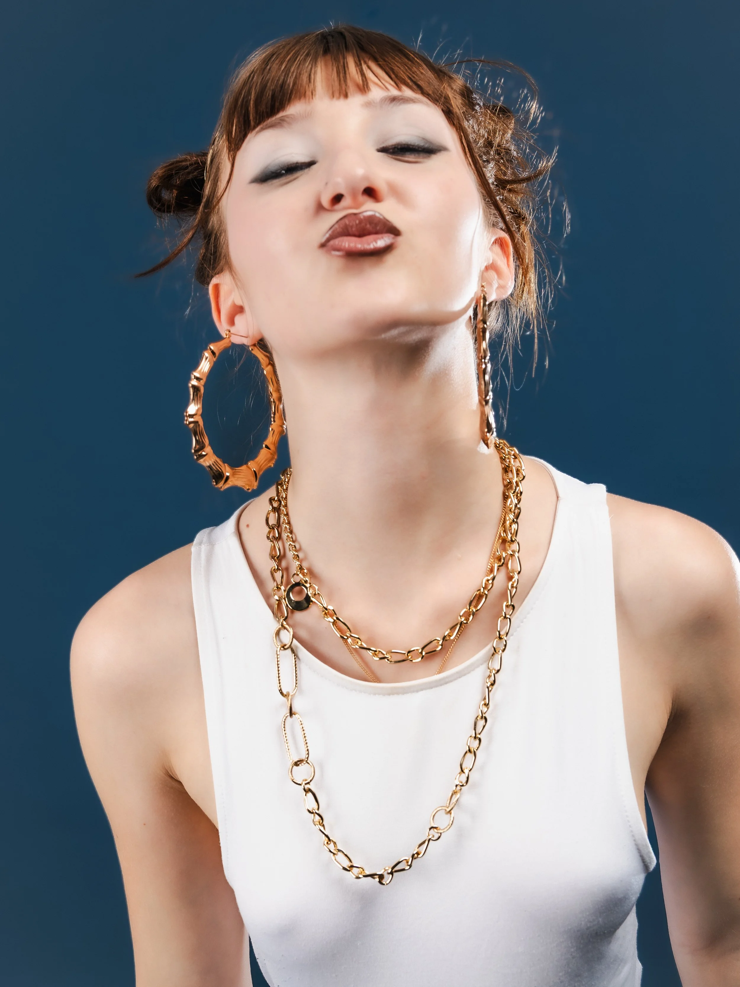 Une femme portant de gros boucles d'oreilles or, une chaîne en or, un débardeur blanc, avec un fond bleu, lui donnant une apparence élégante et confiante.