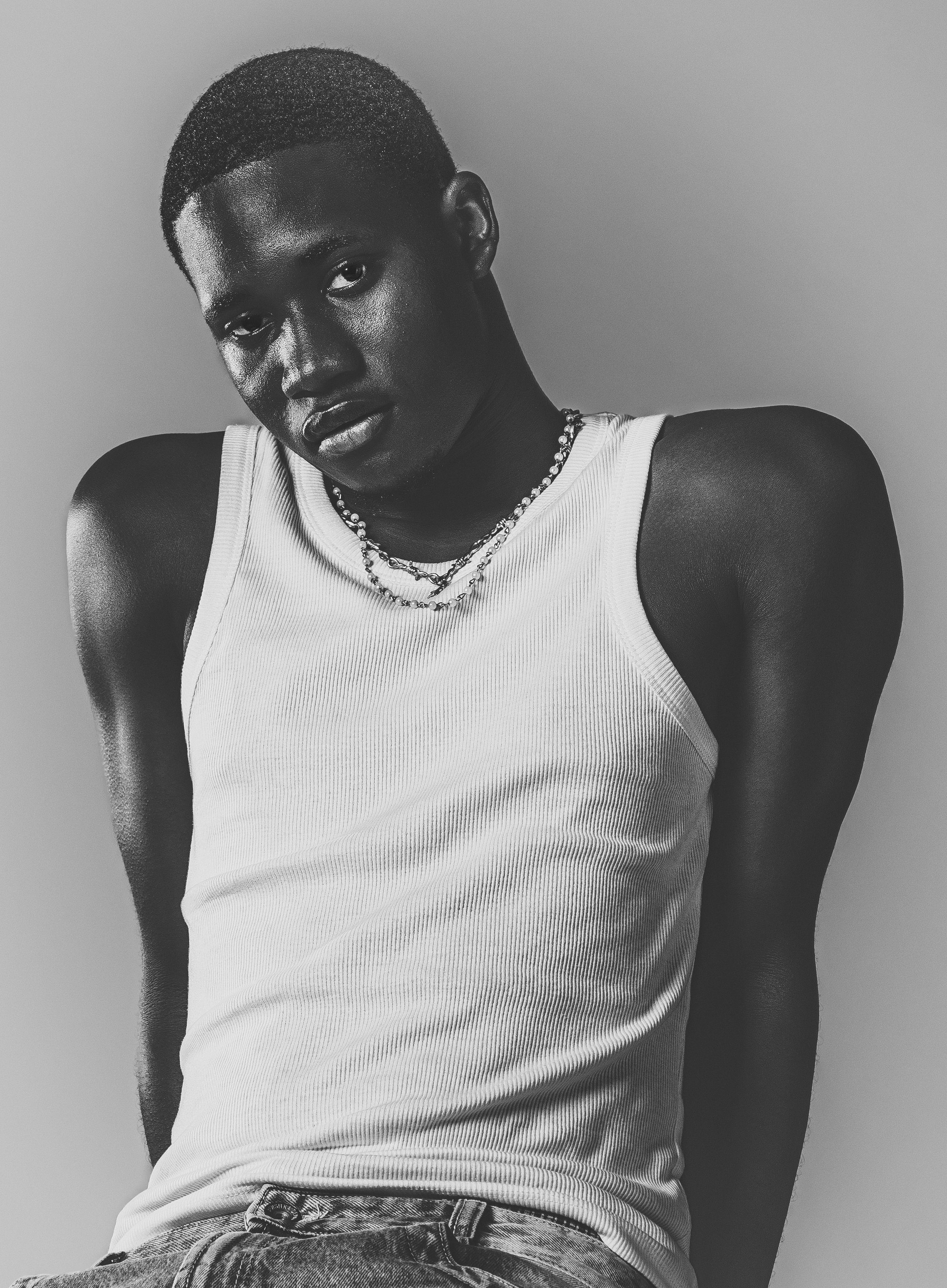 Un jeune homme noir en débardeur blanc, portant des colliers, pose contre un mur gris en noir et blanc.