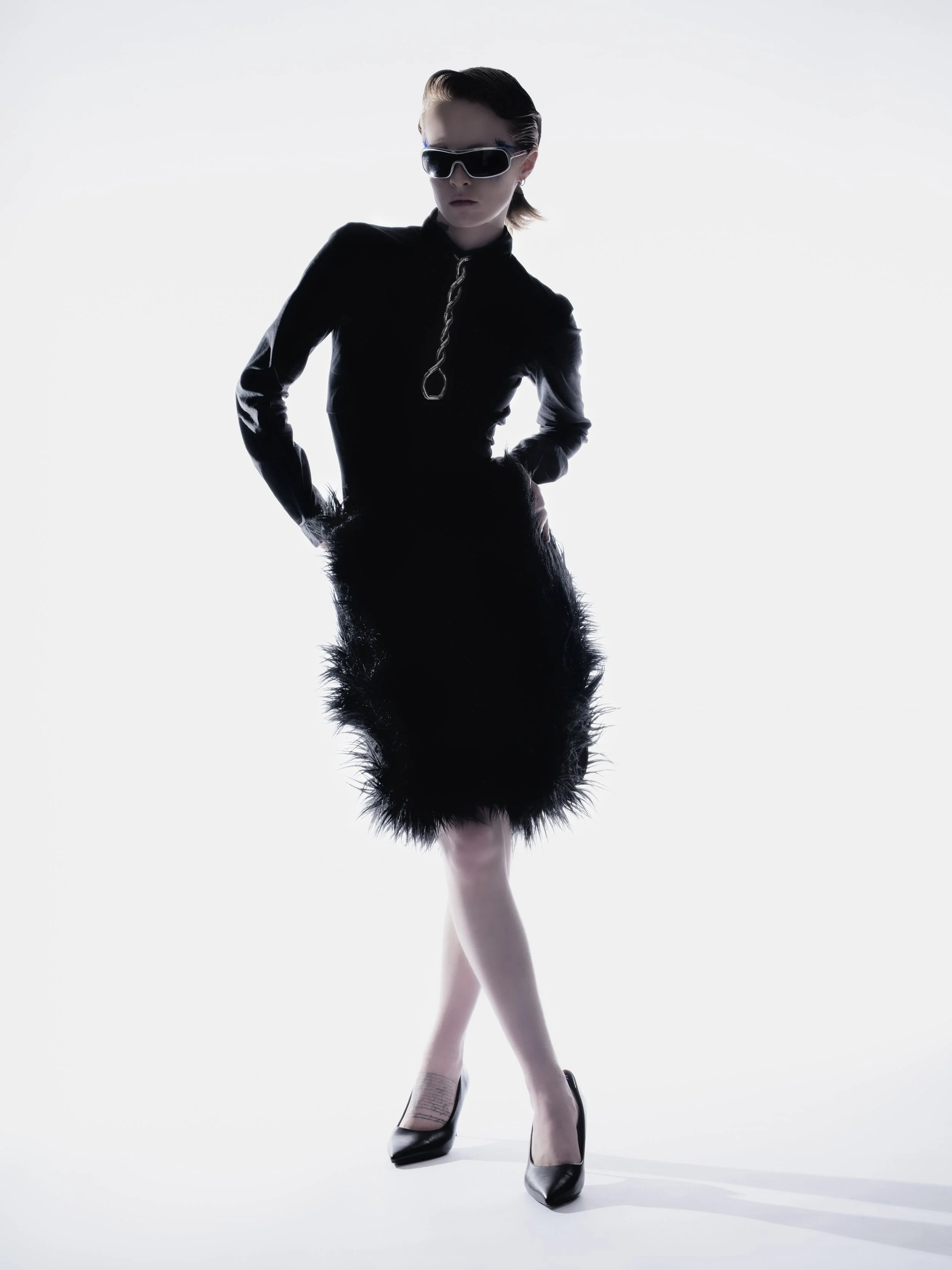 Une femme en silhouette portant une robe noire avec des détails en plumes, des chaussures à talons et des lunettes de soleil dans un fond blanc