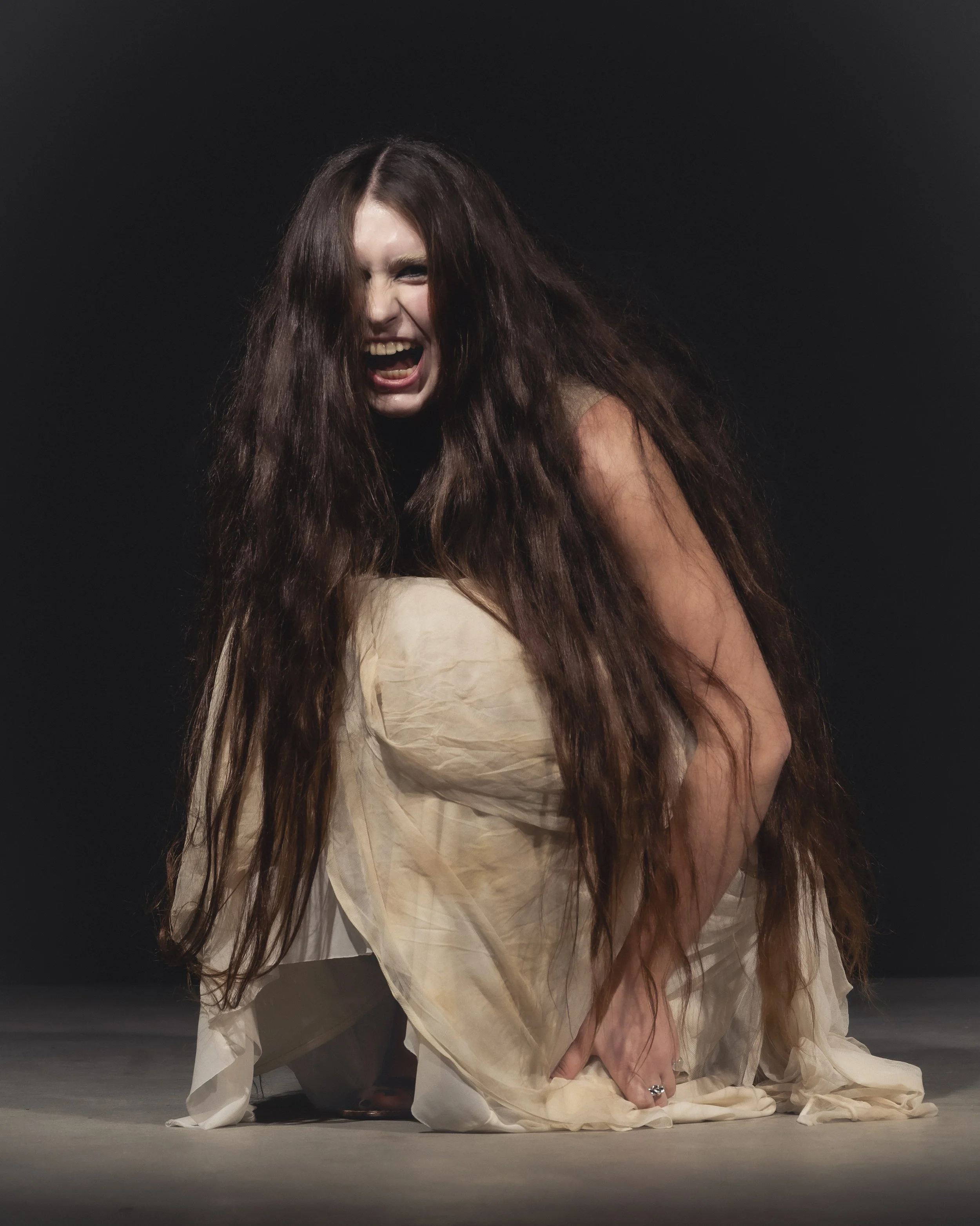 Une femme avec des cheveux longs et bouclés, hurlant ou criant, en position accroupie, portant une robe blanche et un fond noir.