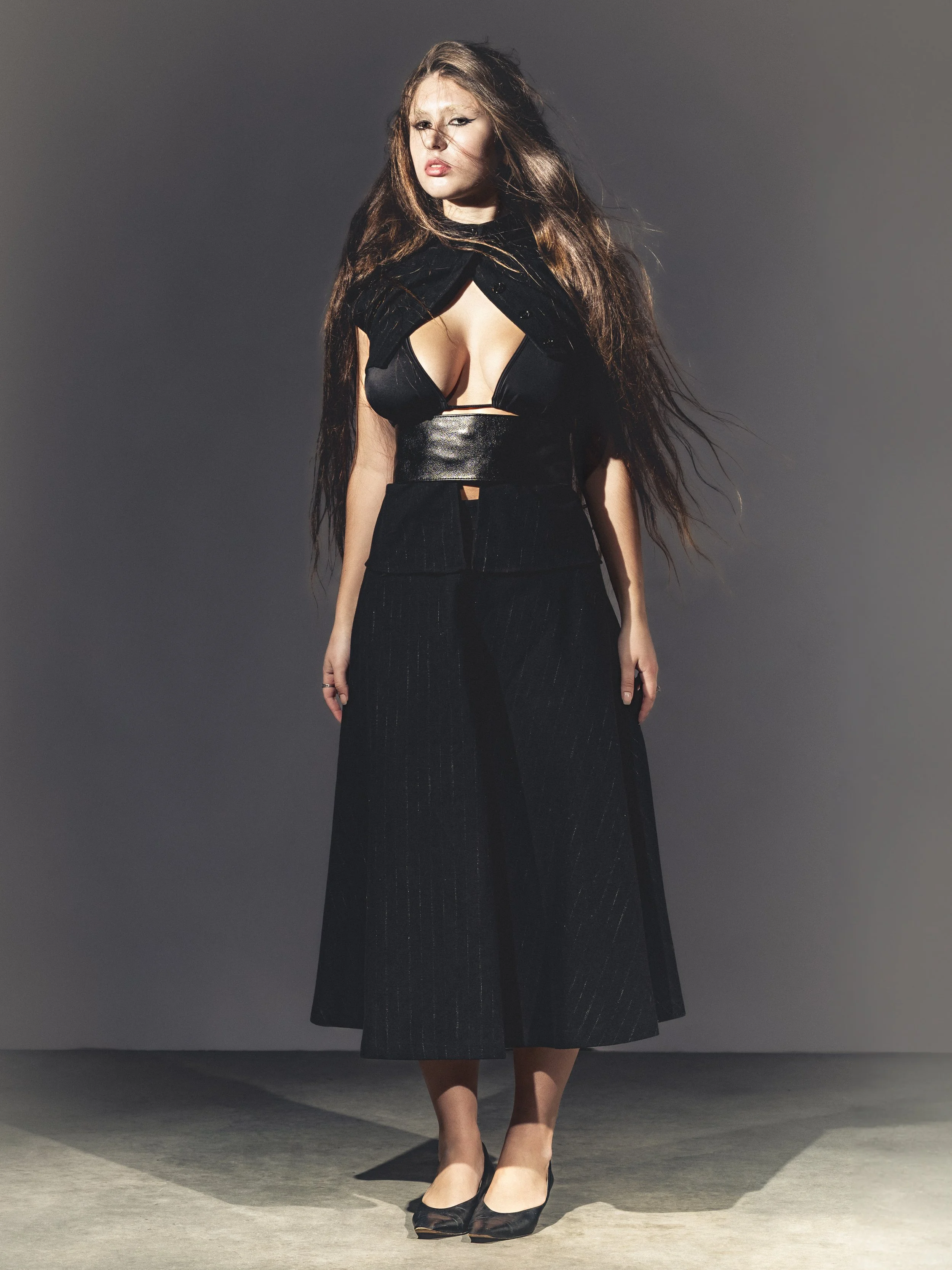 Une femme portant une tenue noire, avec des longues cheveux bruns, se tenant sur un fond gris, regardant vers l'objectif.