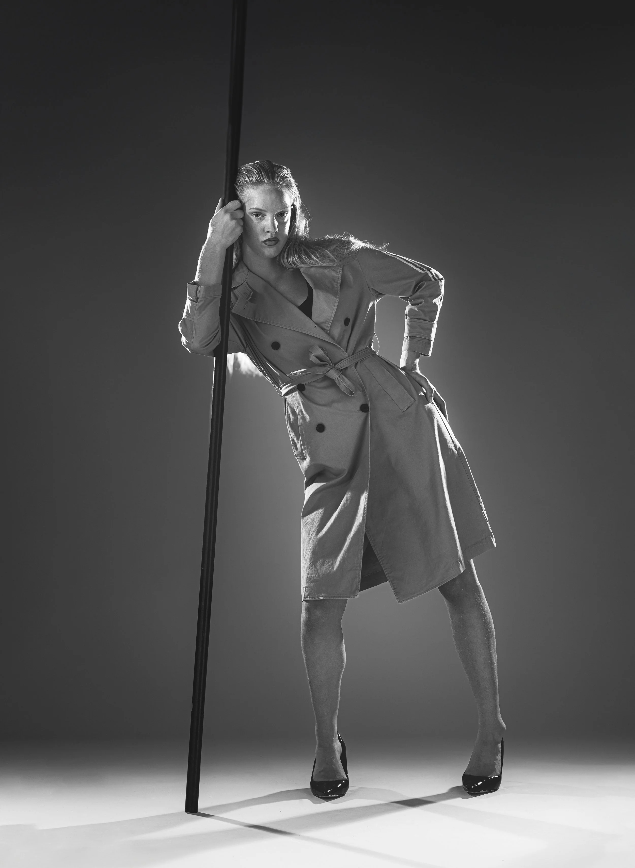 Une femme pose en costume trench coat, portant des talons hauts, dans une séance photo en noir et blanc.