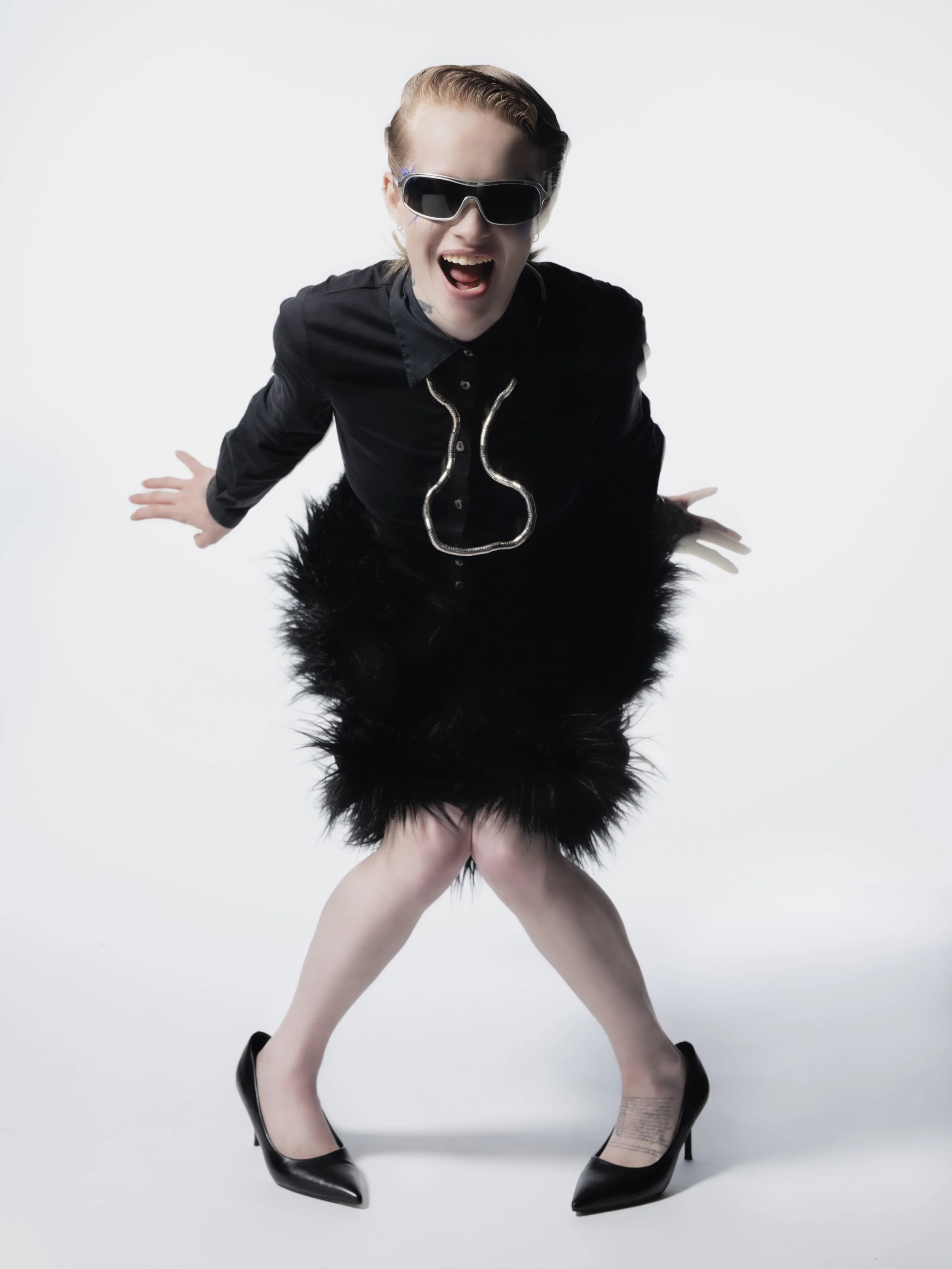 Femme souriante portant des lunettes de soleil, une veste noire élégante, une jupe en plumes noires, et des talons noirs, posant de manière dynamique sur un fond blanc.