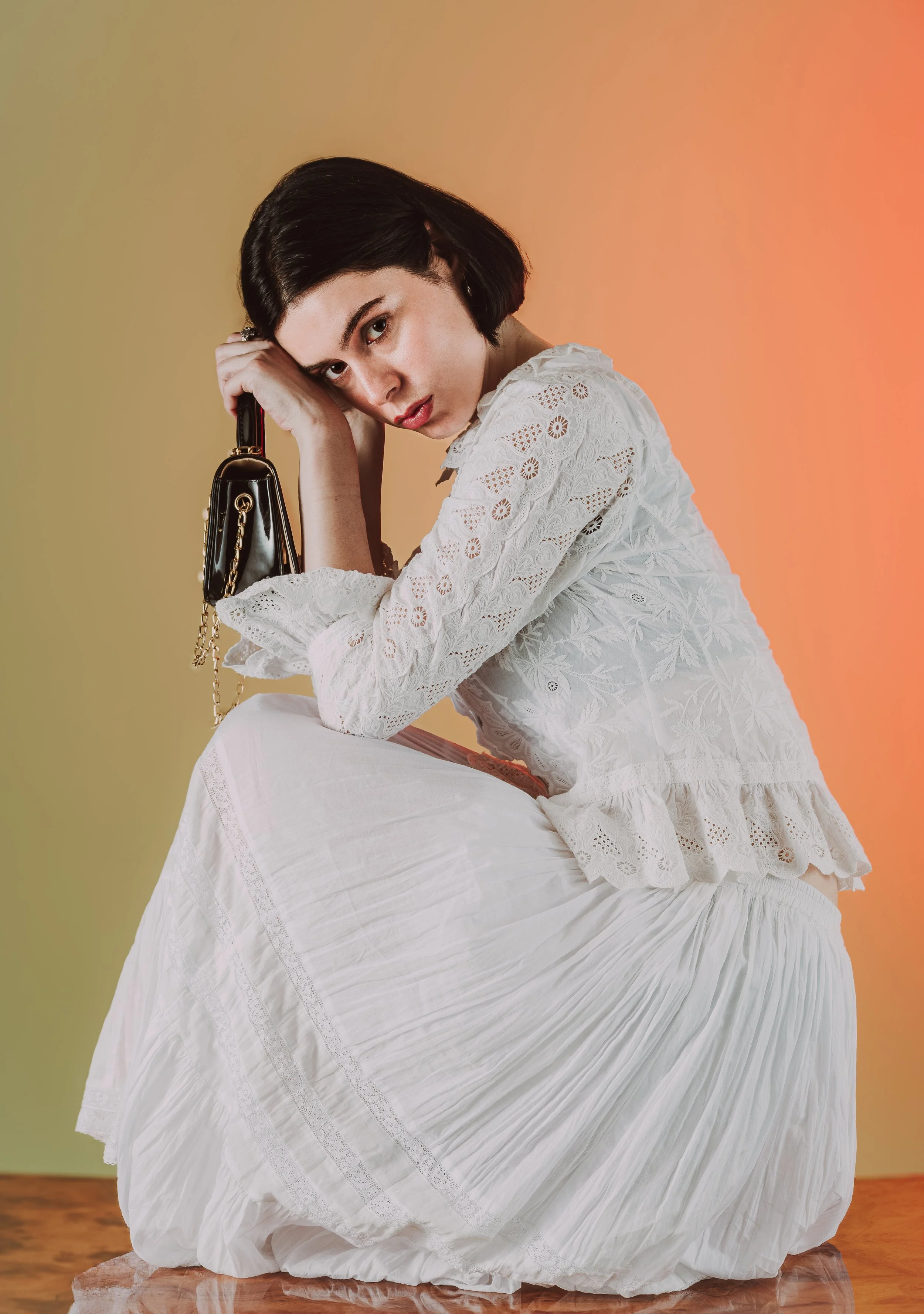 Jeune femme aux cheveux courts, vêtue d'une robe en dentelle blanche, assise sur un sol en bois, tenant un sac à main noir avec chaîne dorée, contre un fond dégradé de couleurs chaudes.
