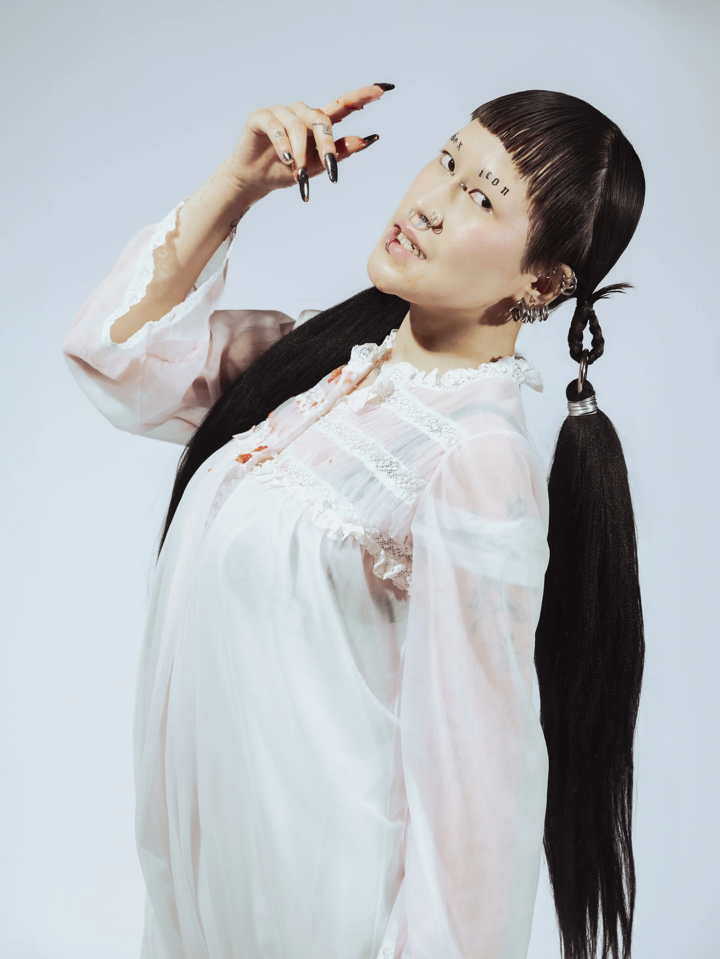 Une personne aux cheveux noirs longs et coiffés en queue de cheval, portant une robe en satin blanc avec des détails en dentelle. Elle a plusieurs piercings au visage, notamment un septum, et porte plusieurs boucles d'oreilles. Elle affiche une expre