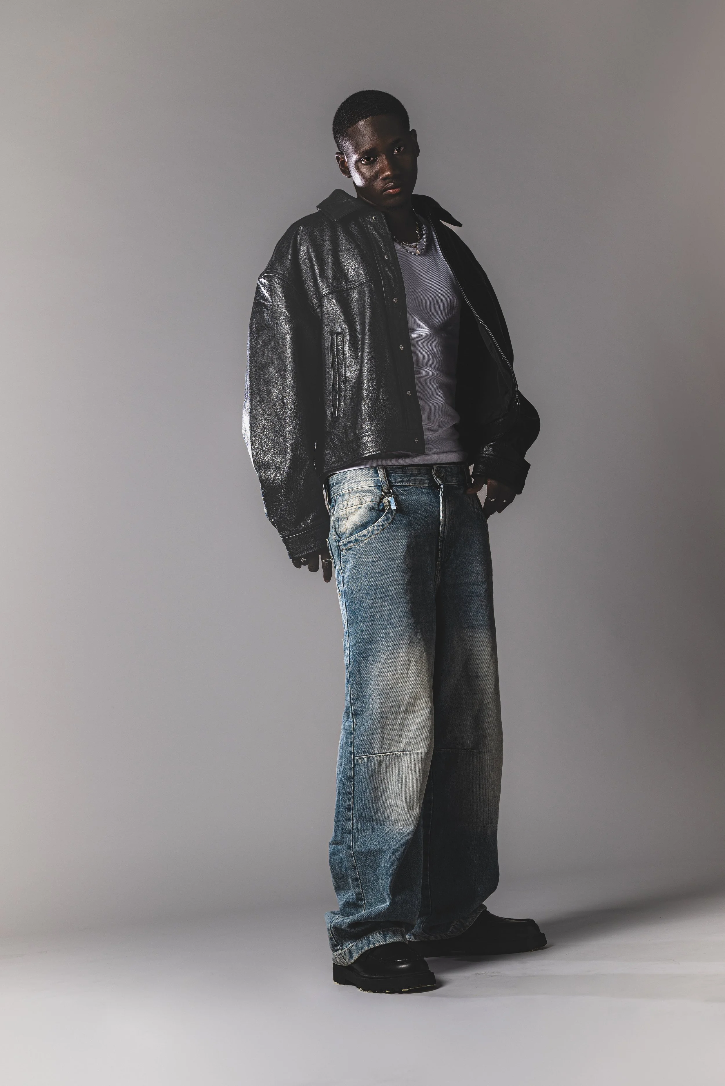 Jeune homme portant une veste en cuir noire, un t-shirt gris, un collier, une paire de jeans délavés et des chaussures noires, debout contre un fond gris.