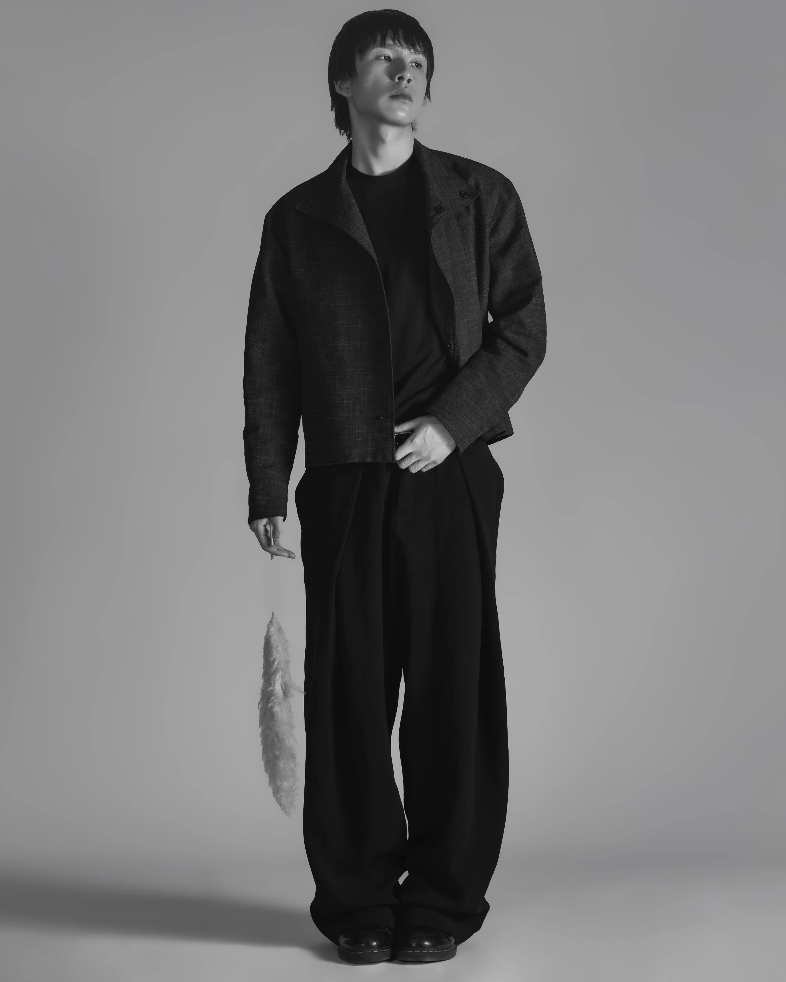 Un jeune homme debout en habits noirs, portant une veste et un pantalon amples, tenant une queue de coq blanche en main, dans une image en noir et blanc.