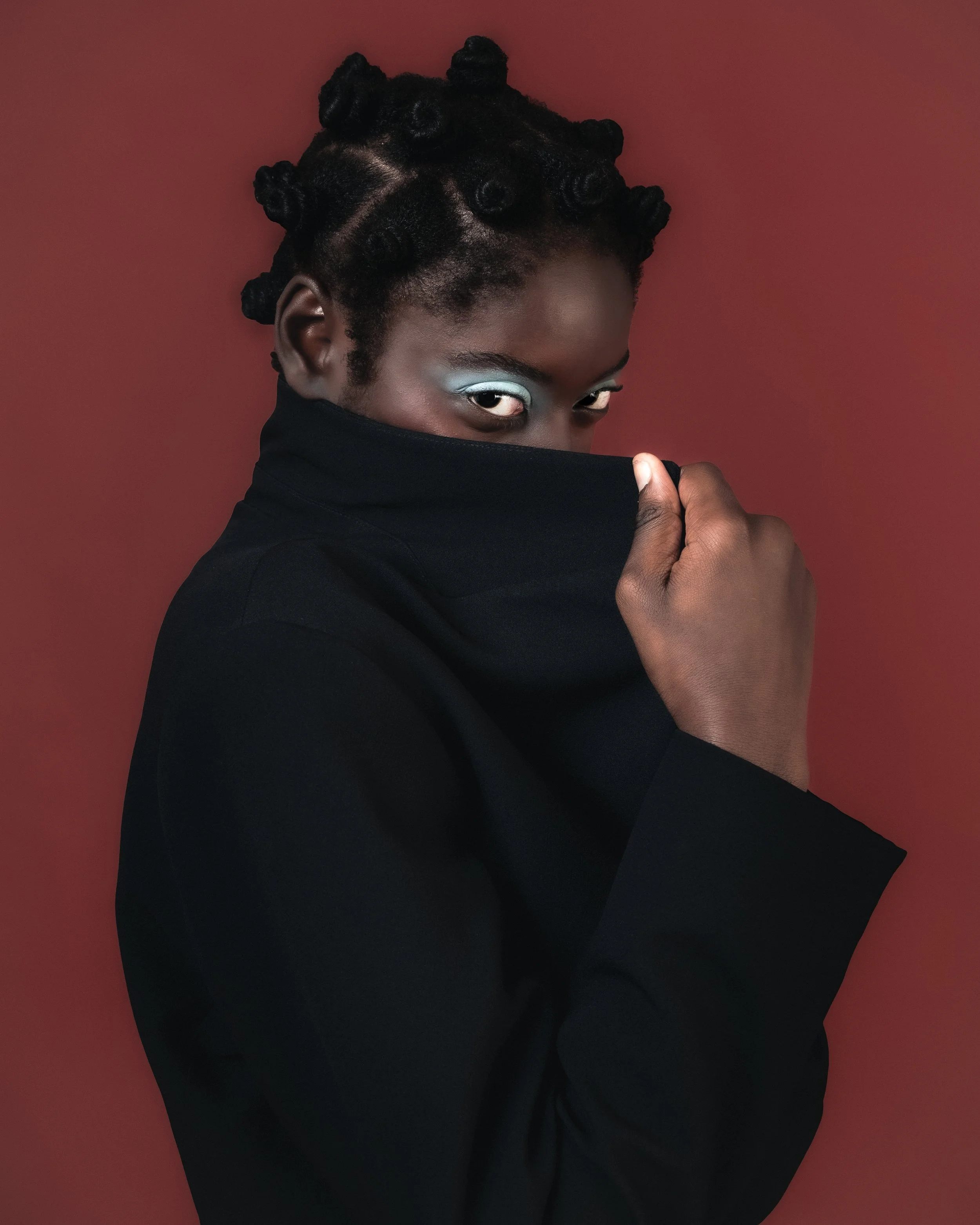 portrait d'une femme noire portant un vêtement noir, couvrant la moitié inférieure du visage, avec un maquillage bleu autour des yeux, contre un fond rouge.