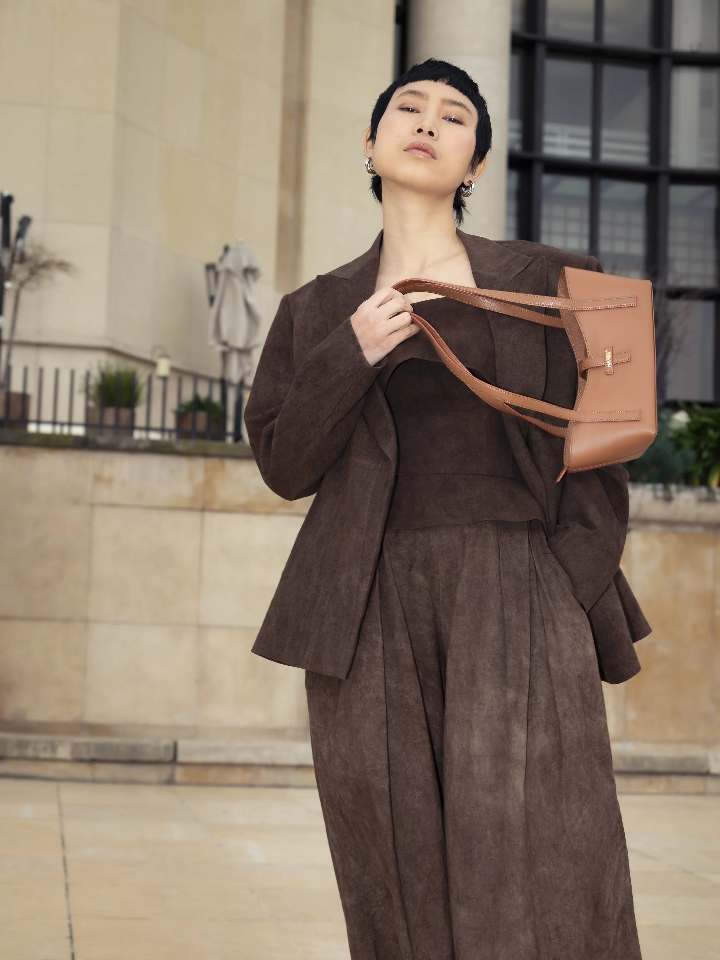 Une femme portant un tailleur marron foncé et tenant un sac beige, debout devant un bâtiment en pierre.
