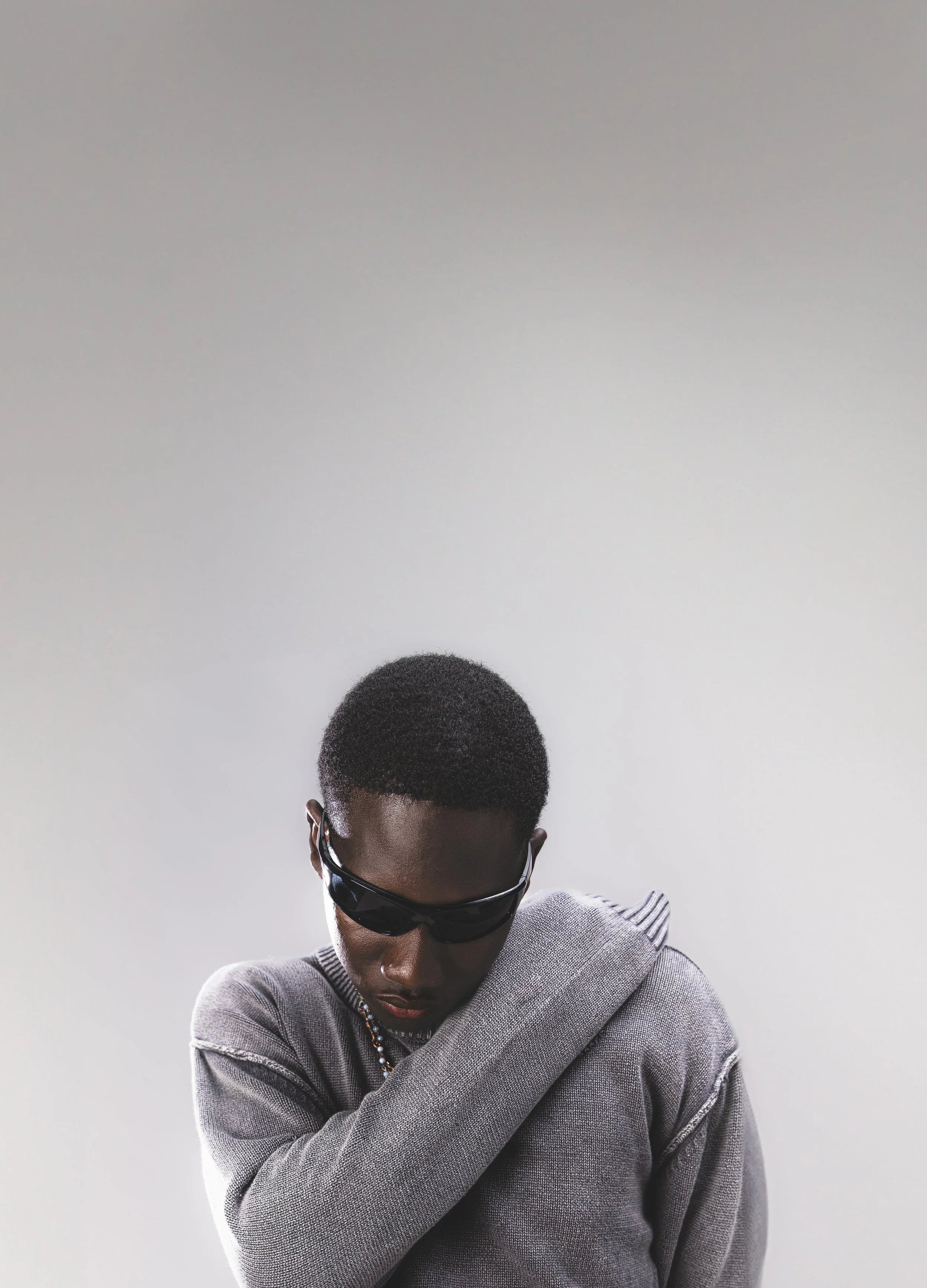 Jeune homme portant des lunettes de soleil noires, en sweat gris, avec un bracelet en perles, contre un fond gris clair.