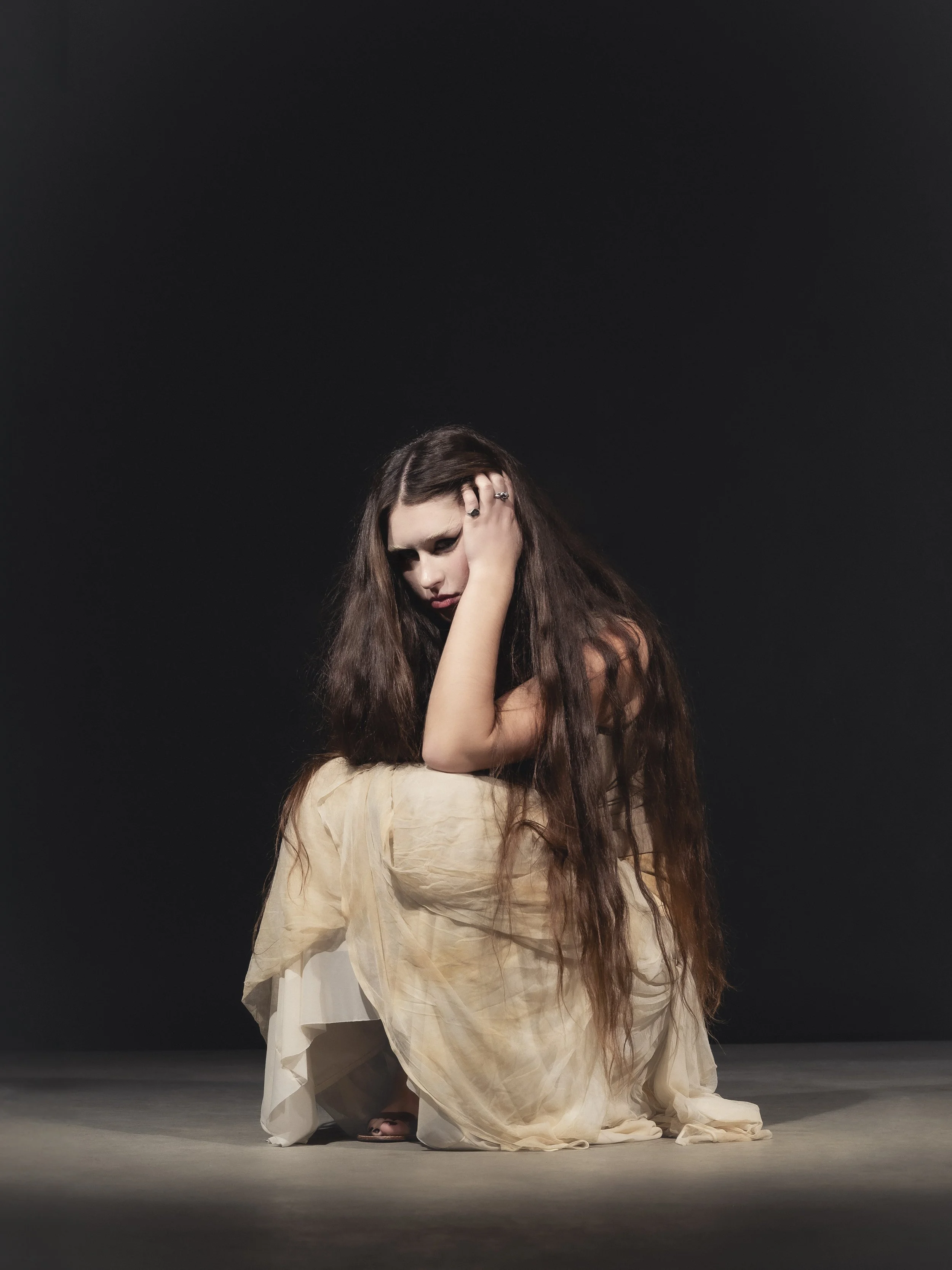 Femme aux longs cheveux bruns portant une robe beige, assise à genoux sur le sol noir, avec une main dans ses cheveux, regardant vers le bas.