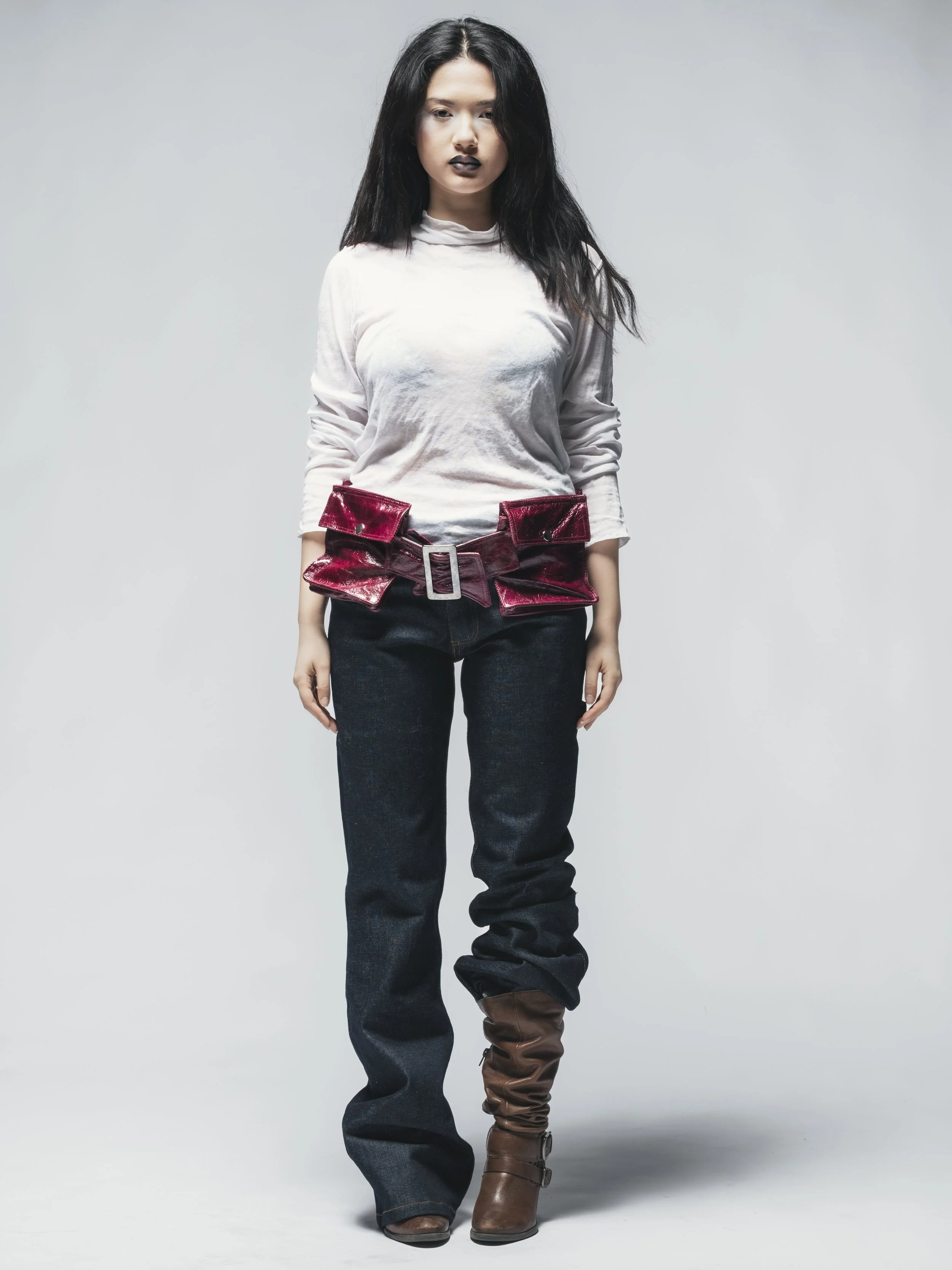 Une femme debout avec des vêtements modernes, portant un haut blanc, un ceinture rouge en velours, un pantalon foncé et des bottes marron.