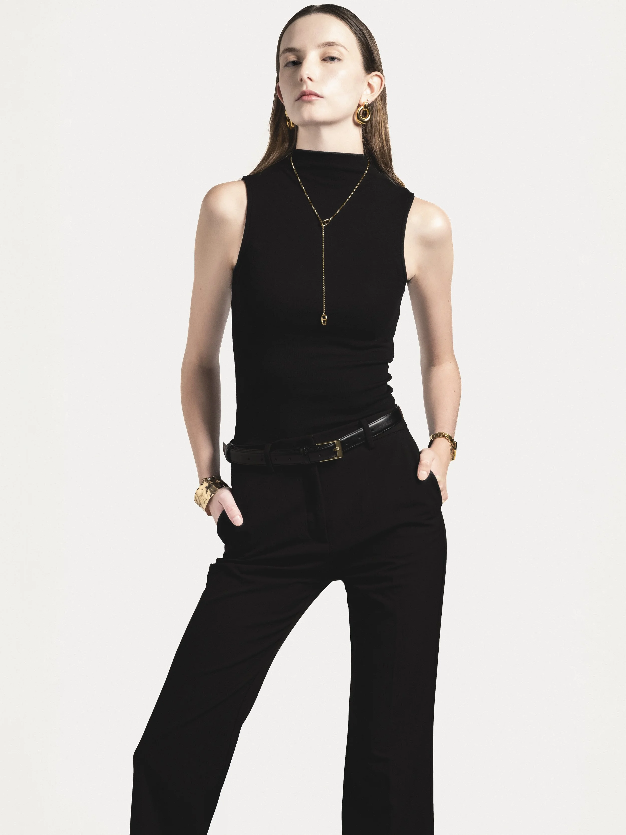 Femme portant un haut noir sans manches, pantalon noir, bijoux en or, contre un fond blanc.
