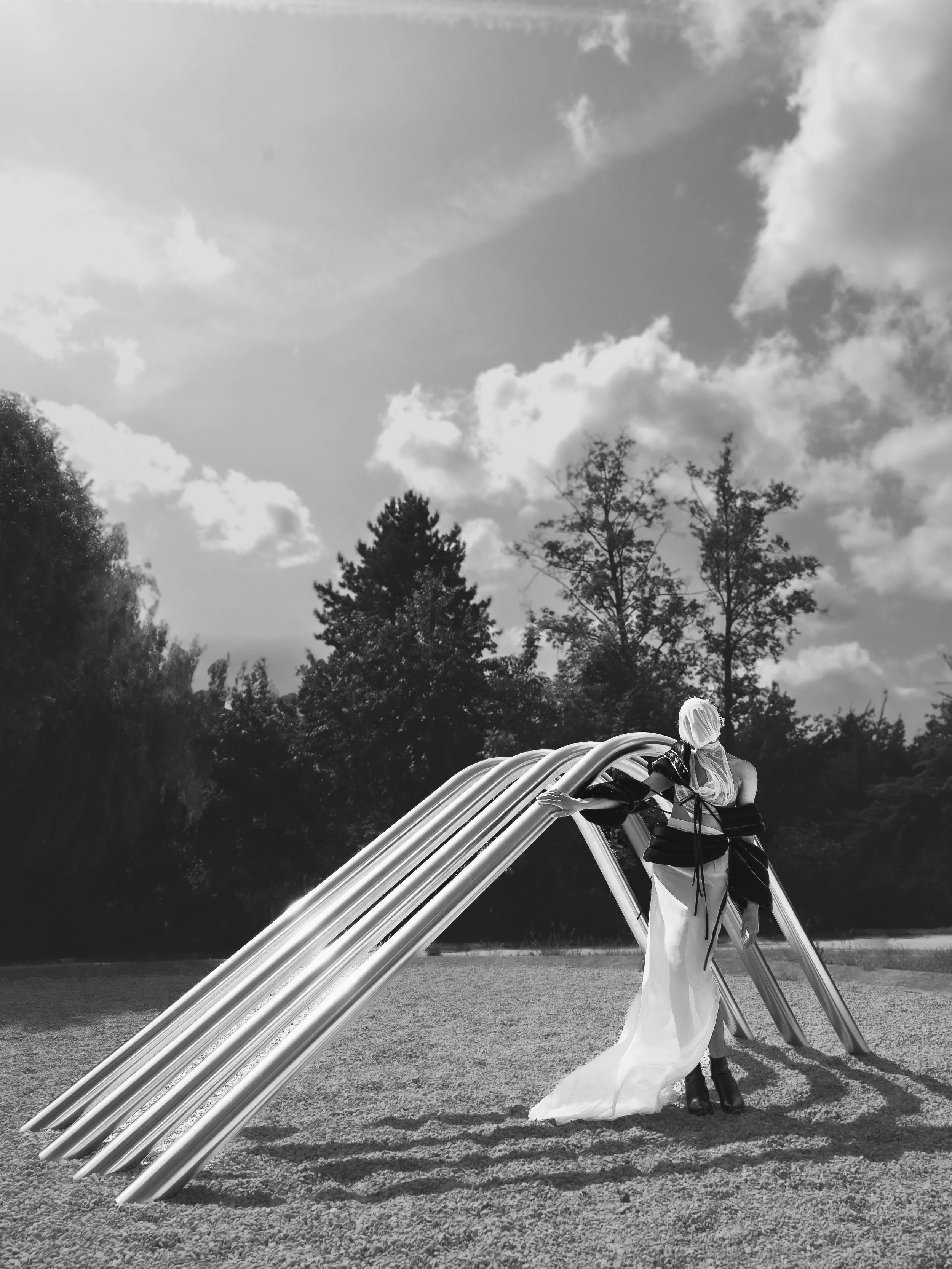 Une femme en robe blanche et voile noir, portant une chaise, se tient dans un parc avec des arbres et un ciel avec des nuages, en noir et blanc.