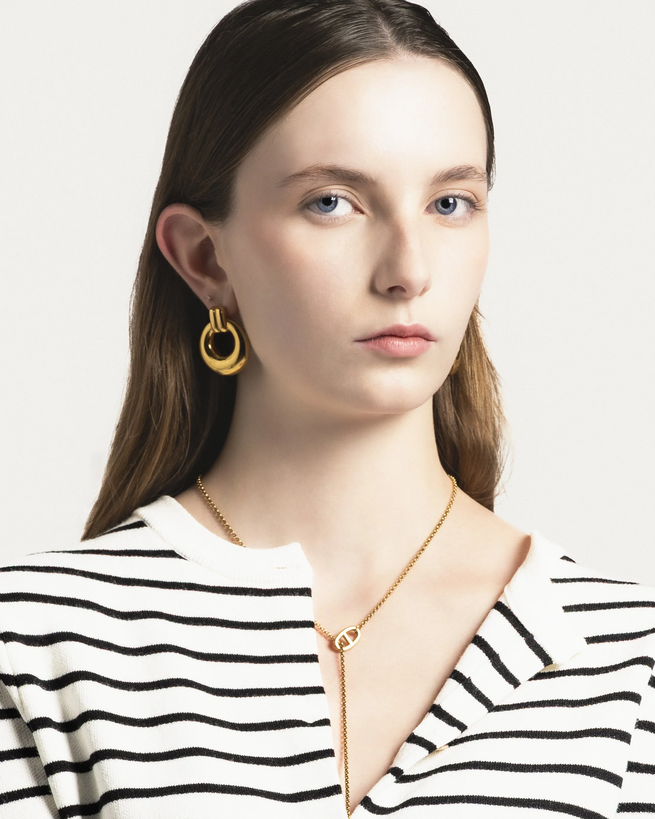 Jeune femme portant des boucles d'oreilles en or, un collier en or et un haut à rayures noires et blanches, avec une expression neutre.