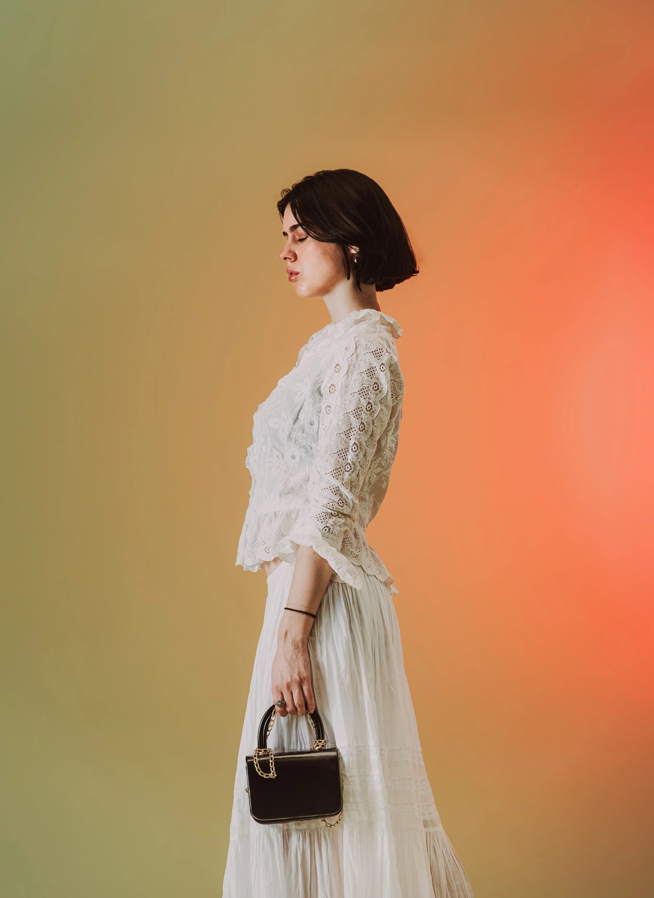 Une femme en robe blanche avec un sac à main noir, de profil, les yeux fermés, contre un fond coloré dégradé.