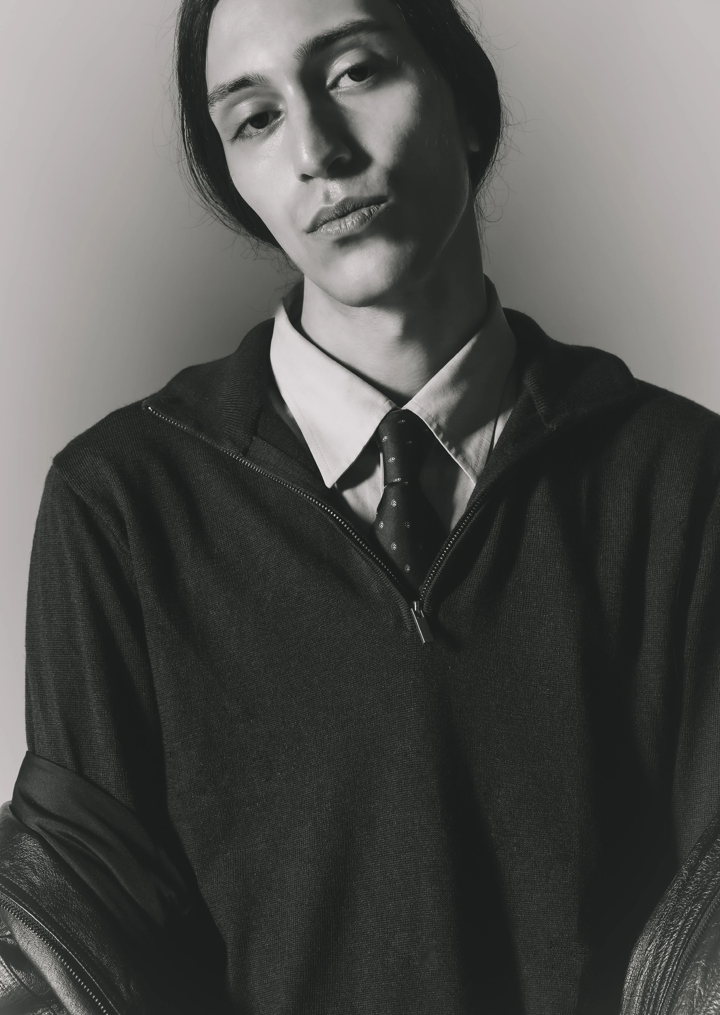 Portrait d'un jeune homme en noir et blanc, portant une chemise, une cravate et une veste à capuche, avec une expression neutre.