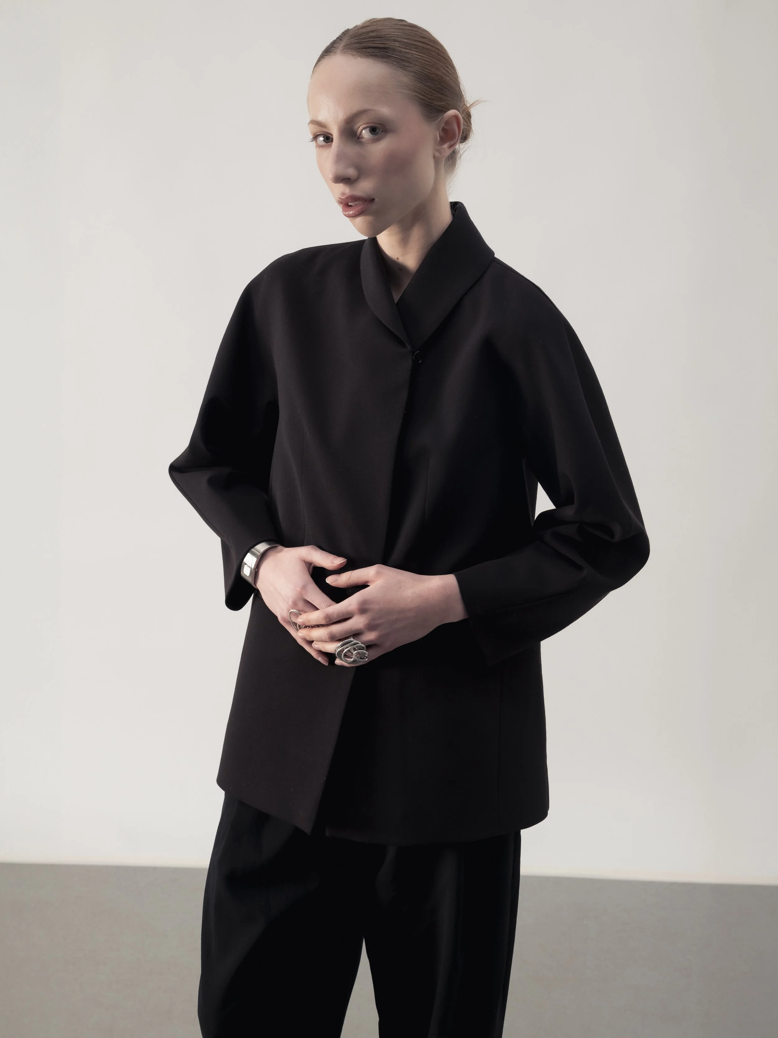 Femme avec cheveux roux attachés, portant un costume noir avec un manteau large, posant devant un fond blanc.