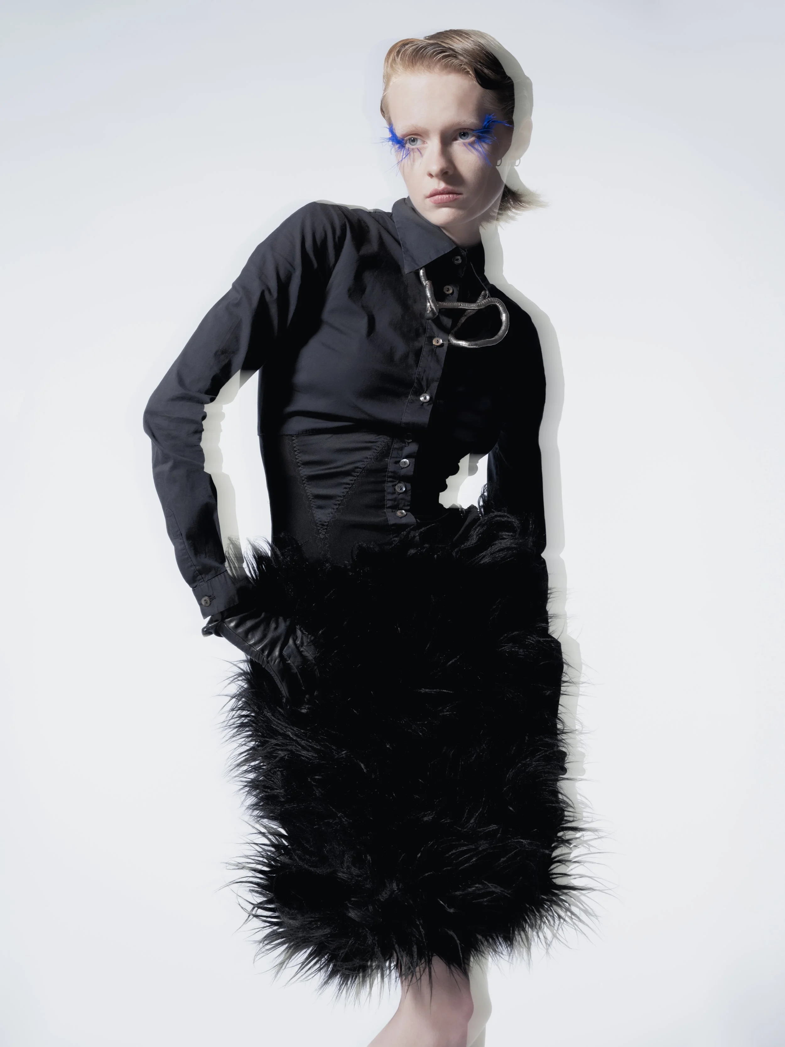 Une femme jeune porte une veste noire, une jupe en fausse fourrure noire, des gants en cuir noirs, avec des yeux accentués de maquillage bleu vif, sur un fond blanc.