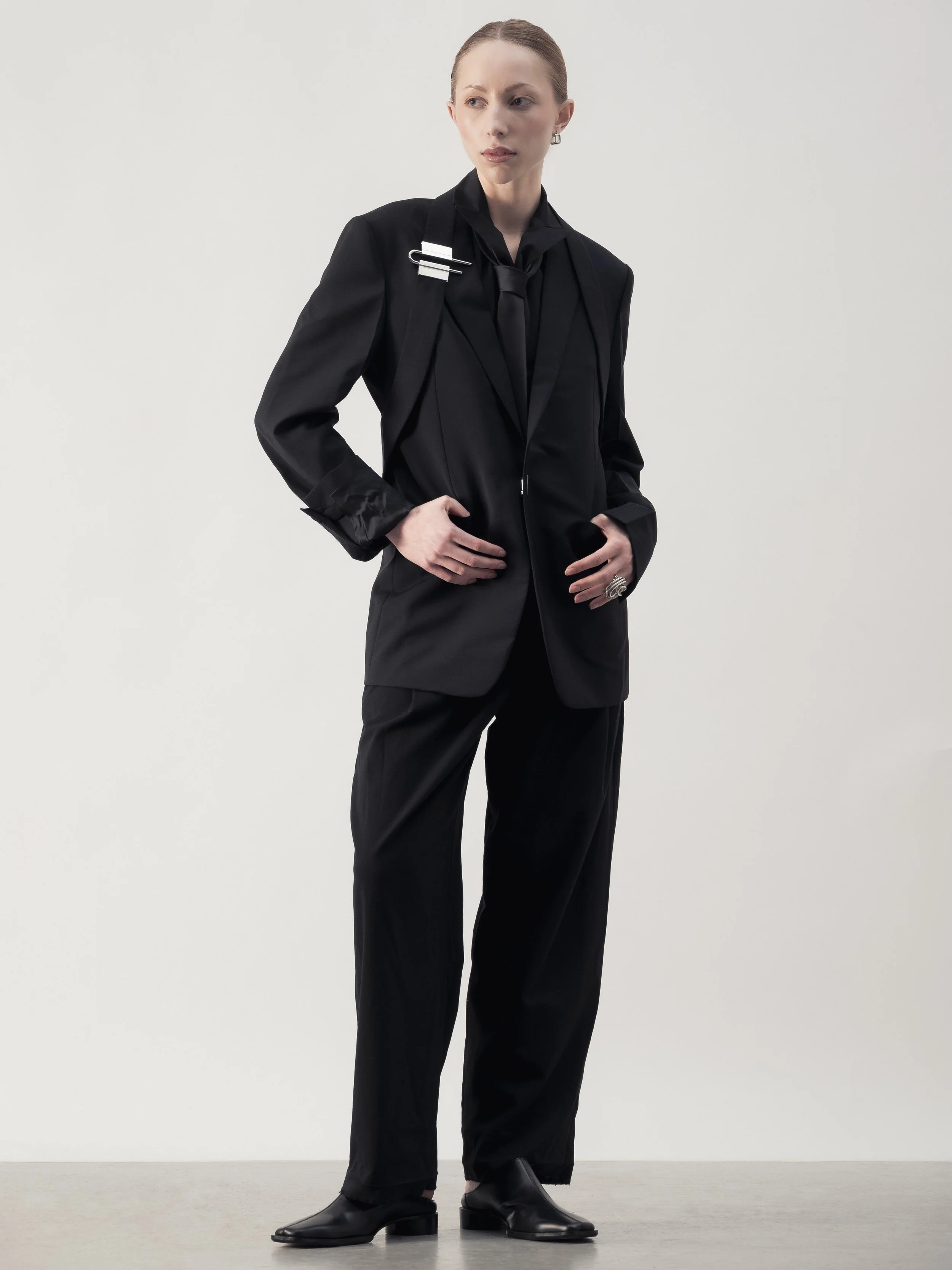 Femme portant un costume noir avec chemise et chaussures en cuir noir dans un fond blanc.