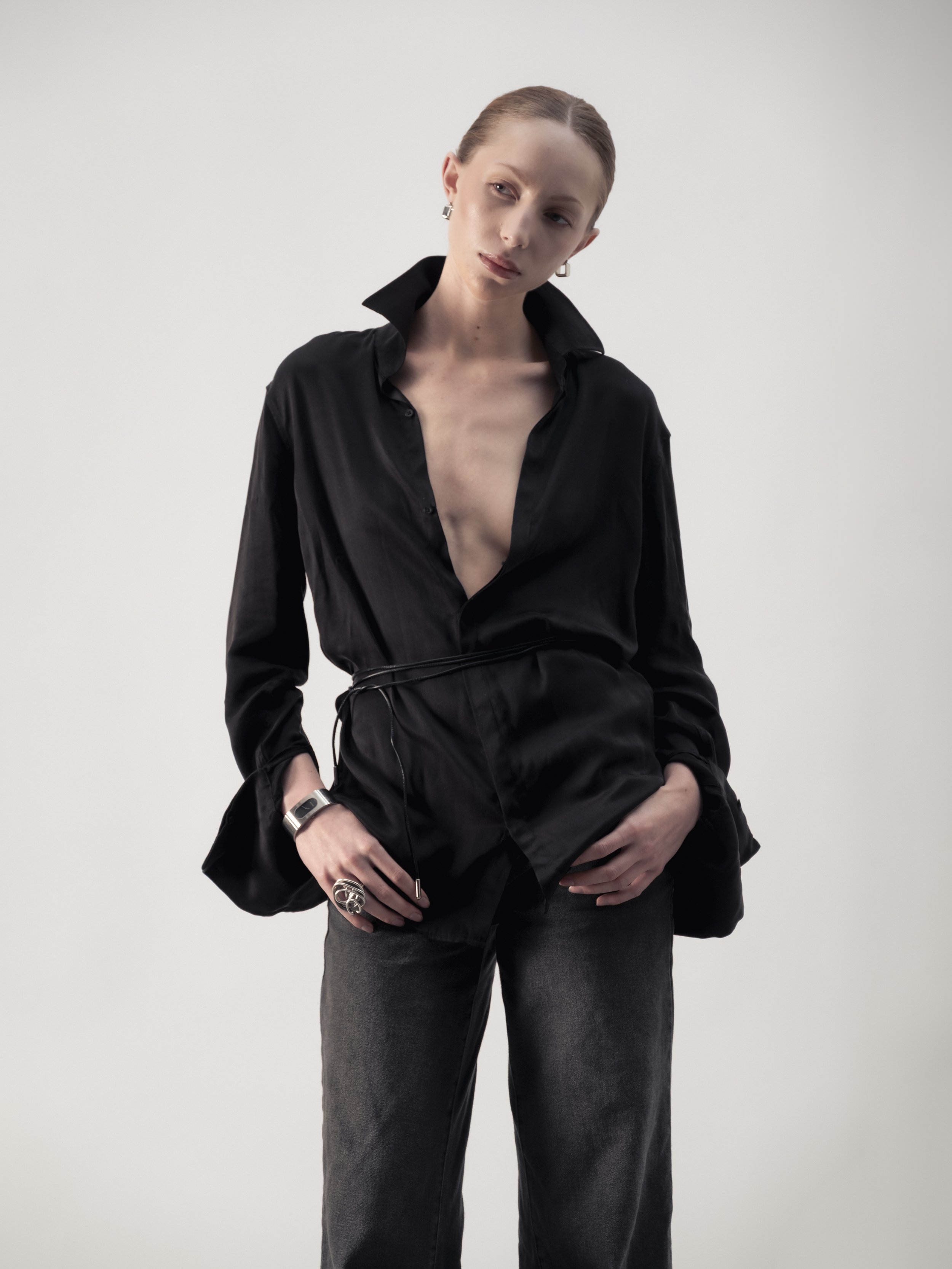 Une femme pose contre un fond blanc, portant une chemise noire décolletée, un pantalon gris et des accessoires en argent.