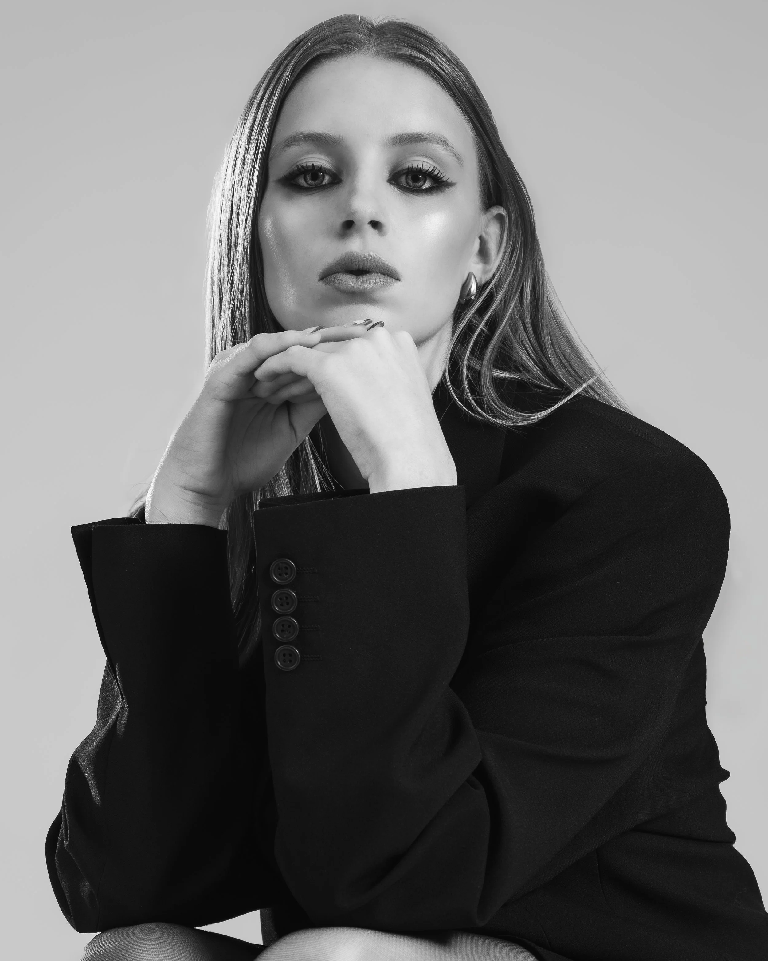 Portrait en noir et blanc d'une femme avec long cheveux, portant un blazer noir, regardant l'objectif, posant avec son menton sur ses mains.