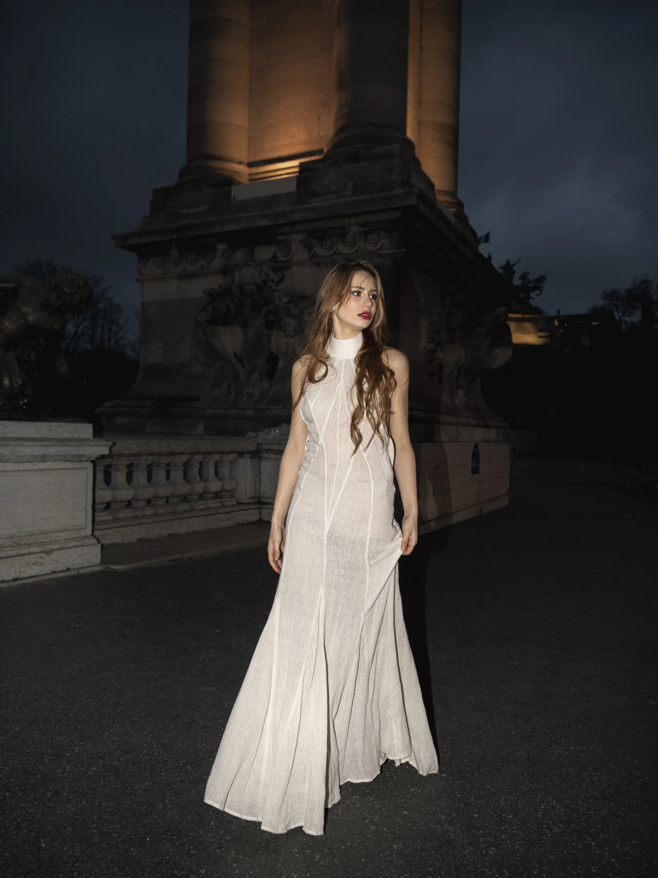 Une femme en robe longue beige se tenant devant une monument historique la nuit.