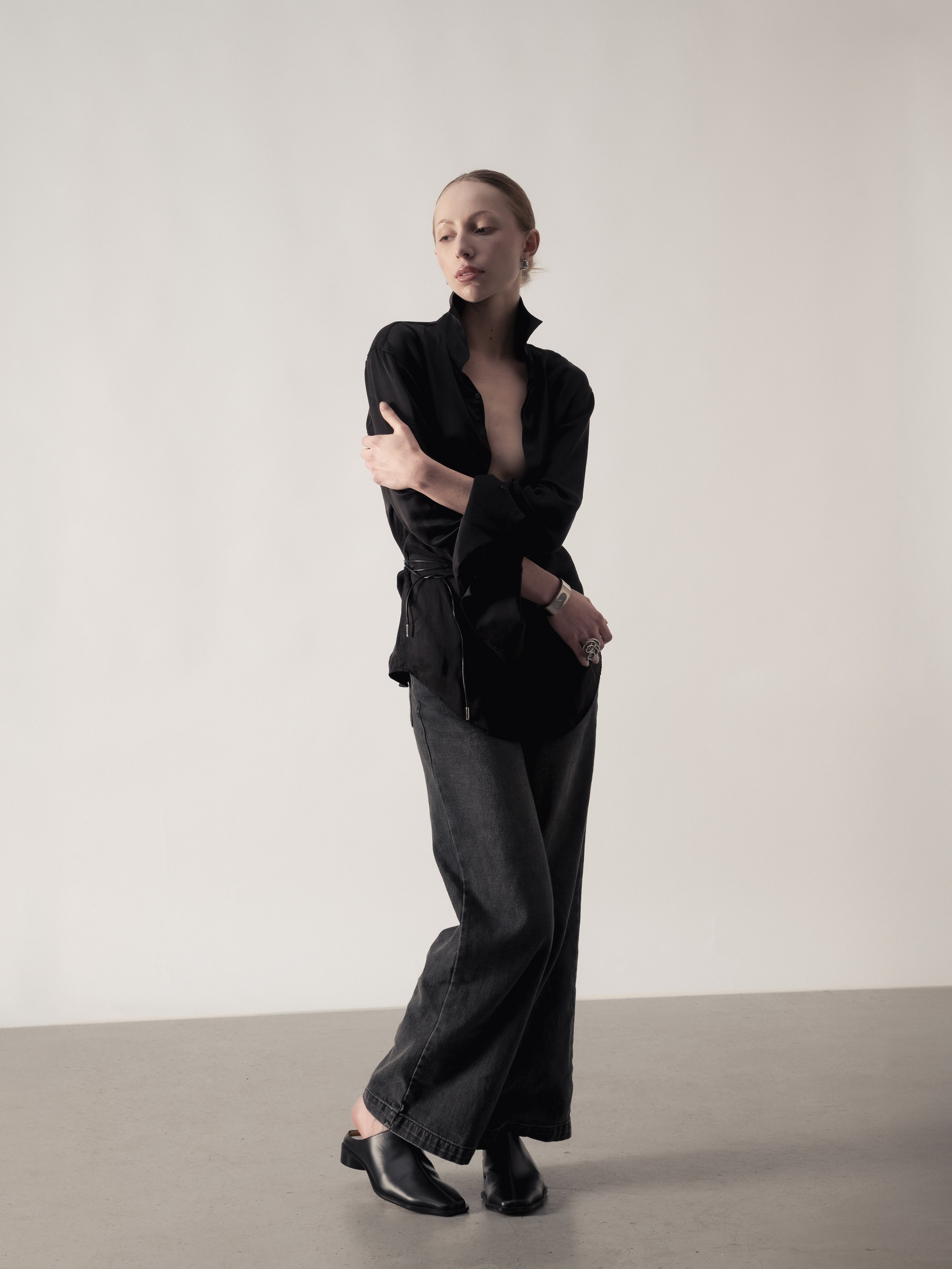 Femme posant dans un studio avec un fond blanc, portant un haut noir, un pantalon gris large et des bottines noires.