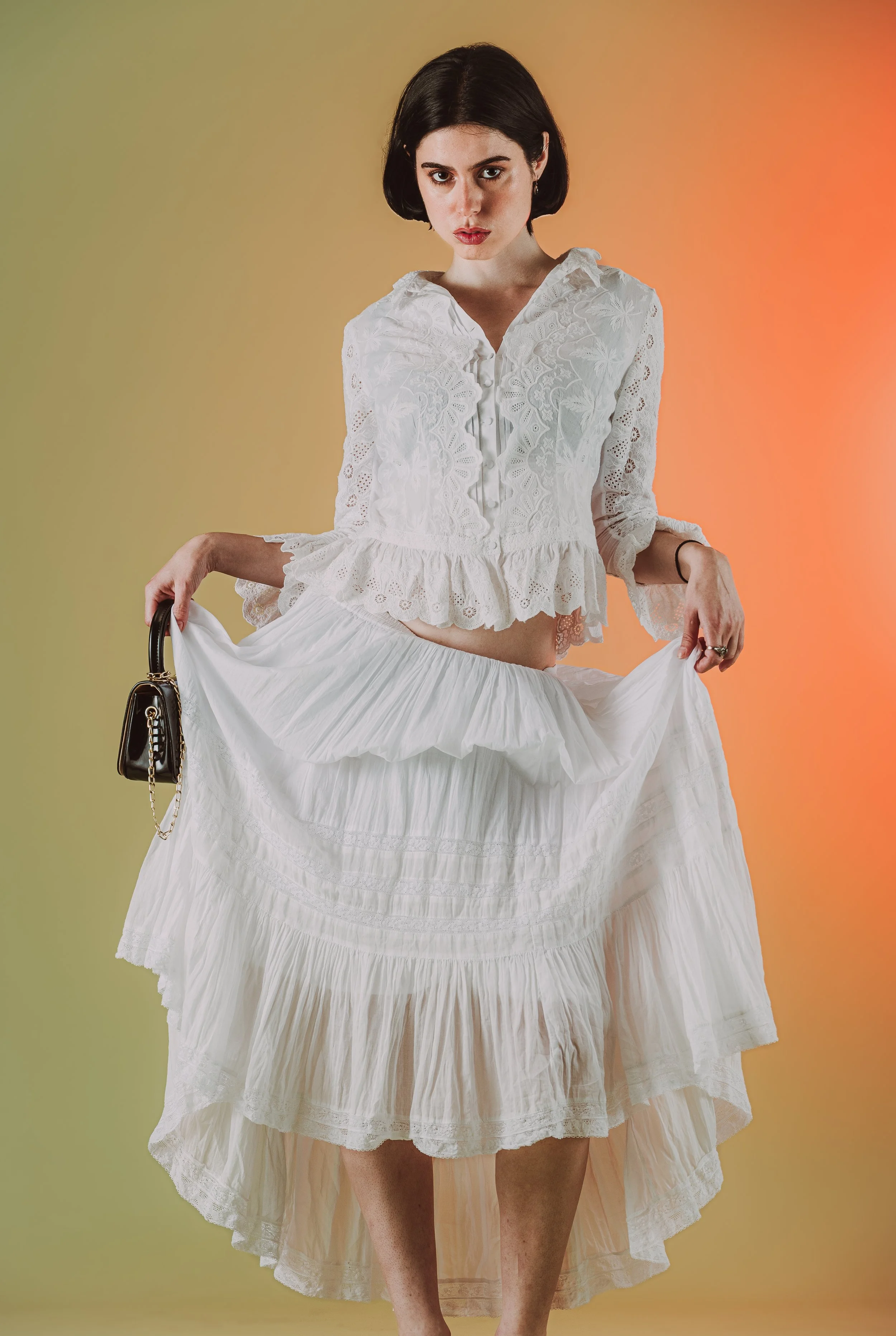 Une femme portant une robe longue blanche avec une chemise en dentelle et un jupon, tenant un petit sac noir avec chaîne