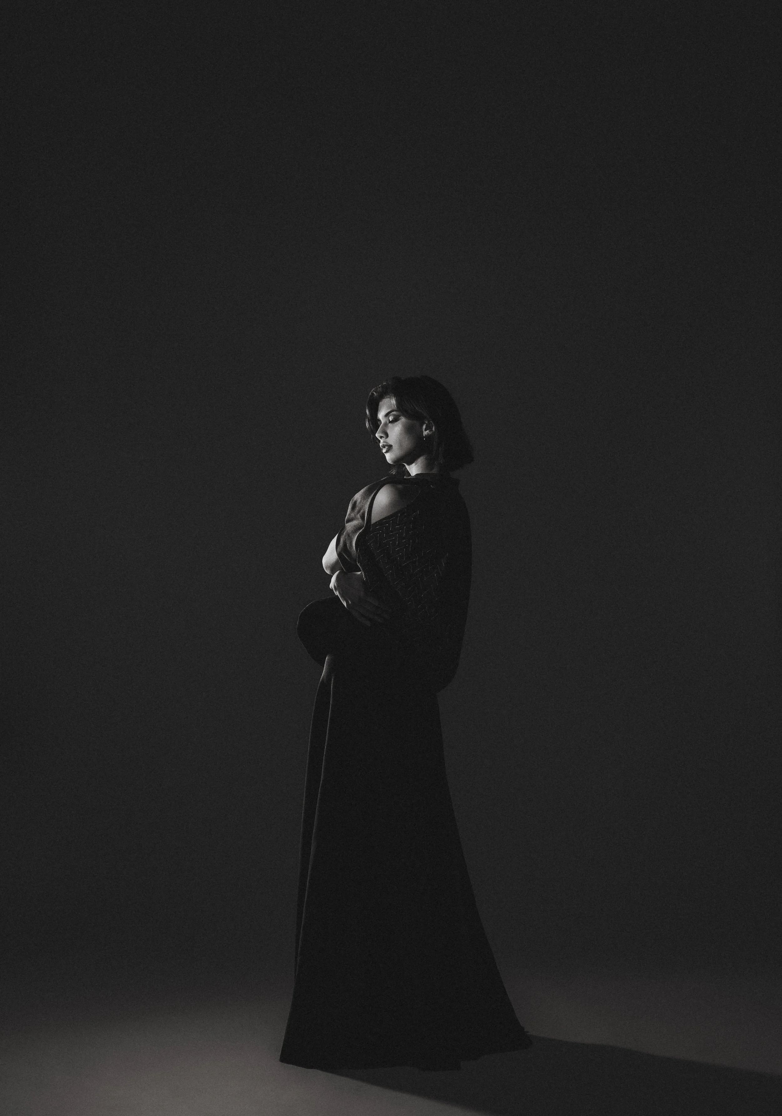 Une femme en robe longue noire, de profil, dans un décor sombre, éclairée par une lumière douce, en noir et blanc.