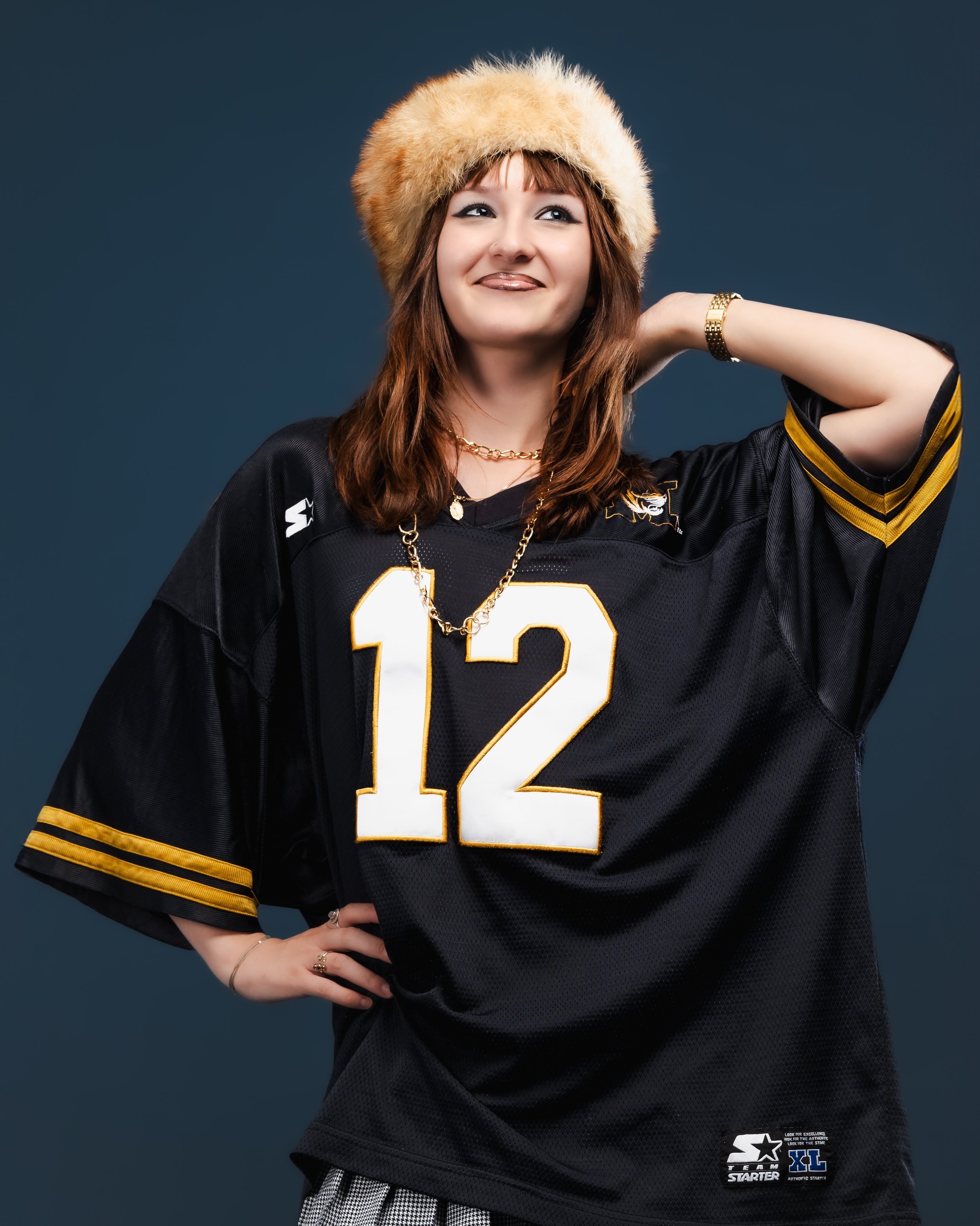 Jeune femme souriante portant un maillot de football noir avec le numéro 12, un chapeau en fourrure beige, accessoires en or, et posant avec la main sur la hanche contre un fond bleu.