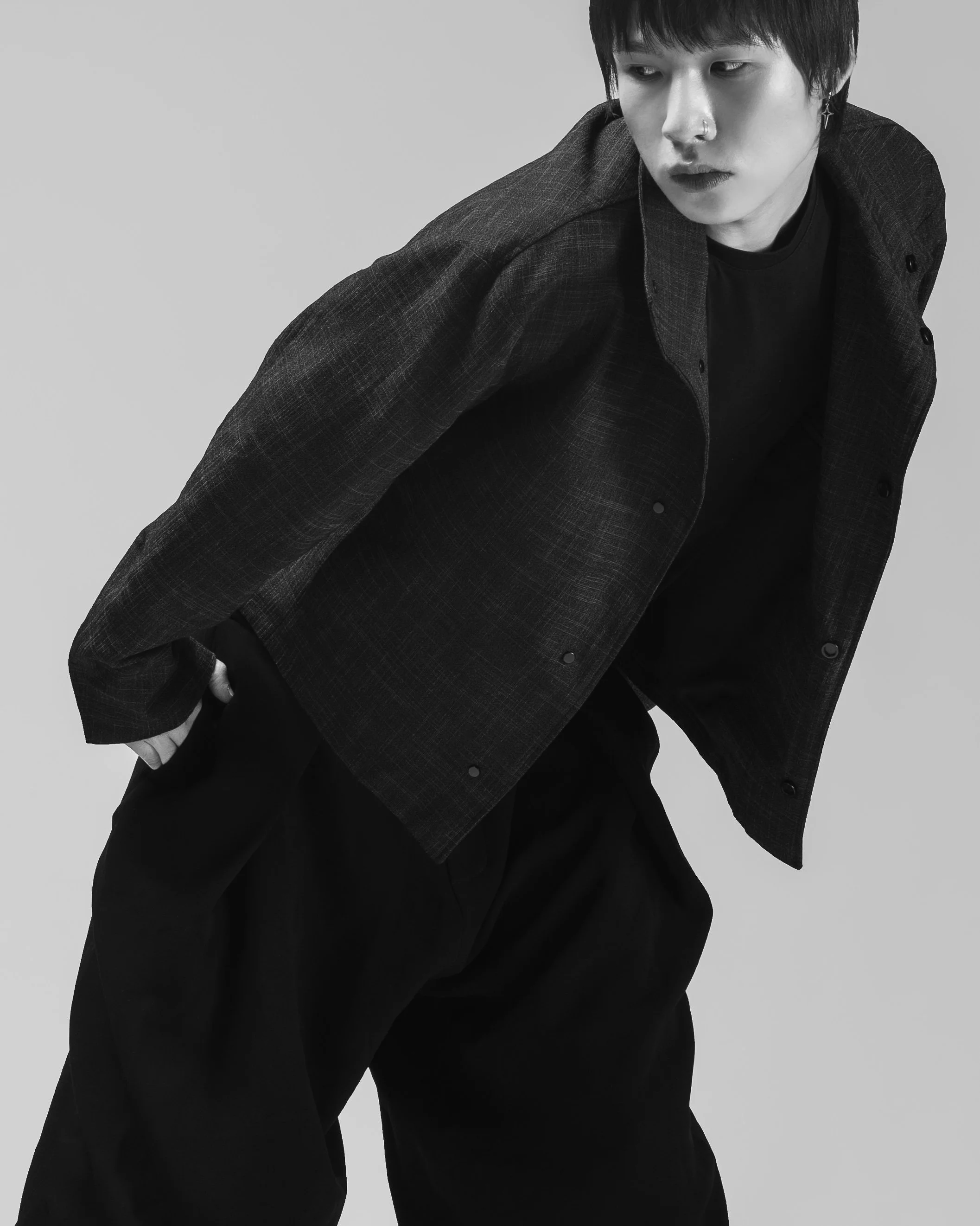 Un jeune homme portant un manteau foncé et un pantalon ample pose pour une séance photo en noir et blanc contre un fond clair.