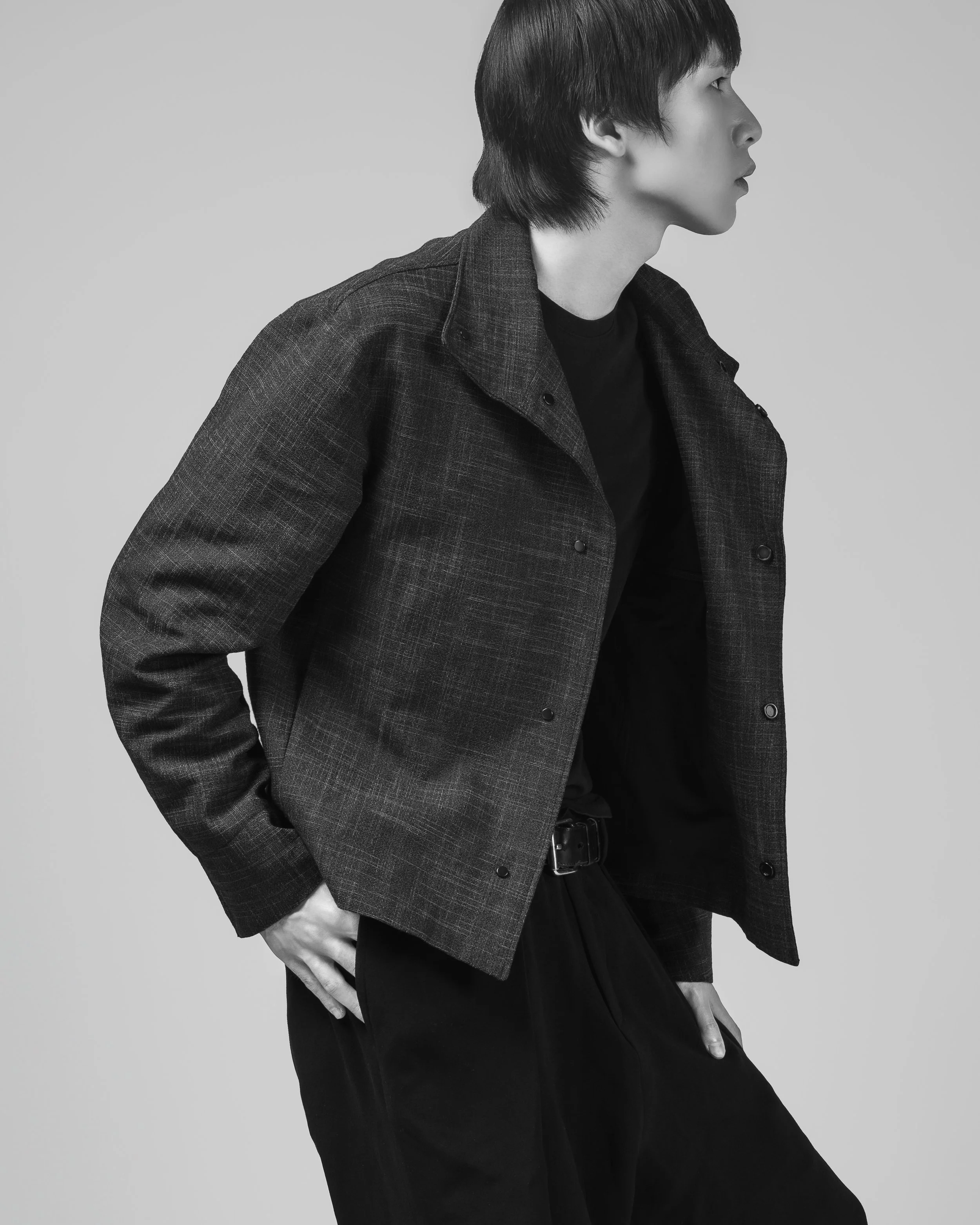 Une personne au profil, portant une veste en tissu à carreaux et un pantalon noir, avec une main dans la poche, dans un style monochrome.