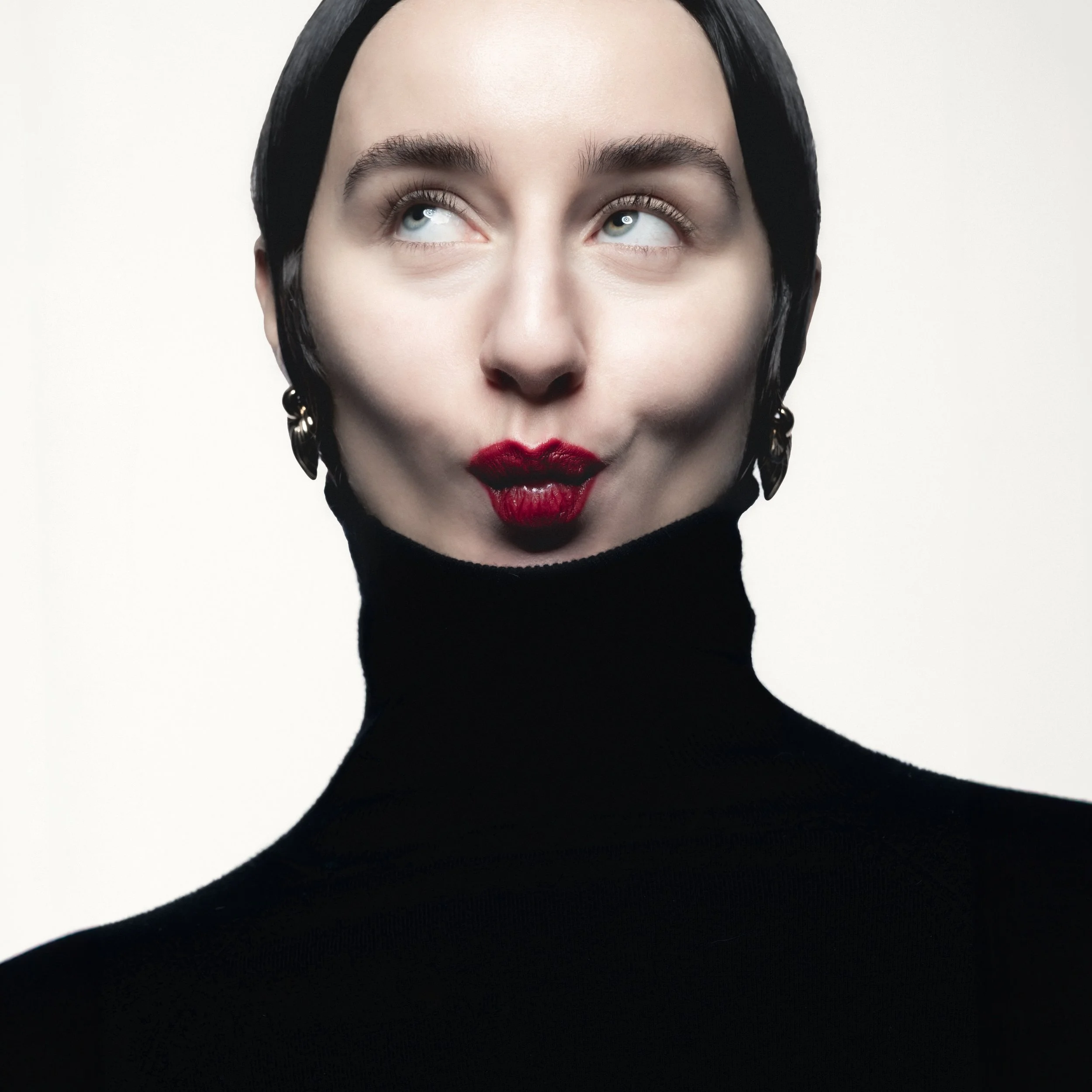 Portrait d'une femme avec une expression amusée, portant un col roulé noir et des boucles d'oreilles, avec des lèvres rouges et des yeux bleus.