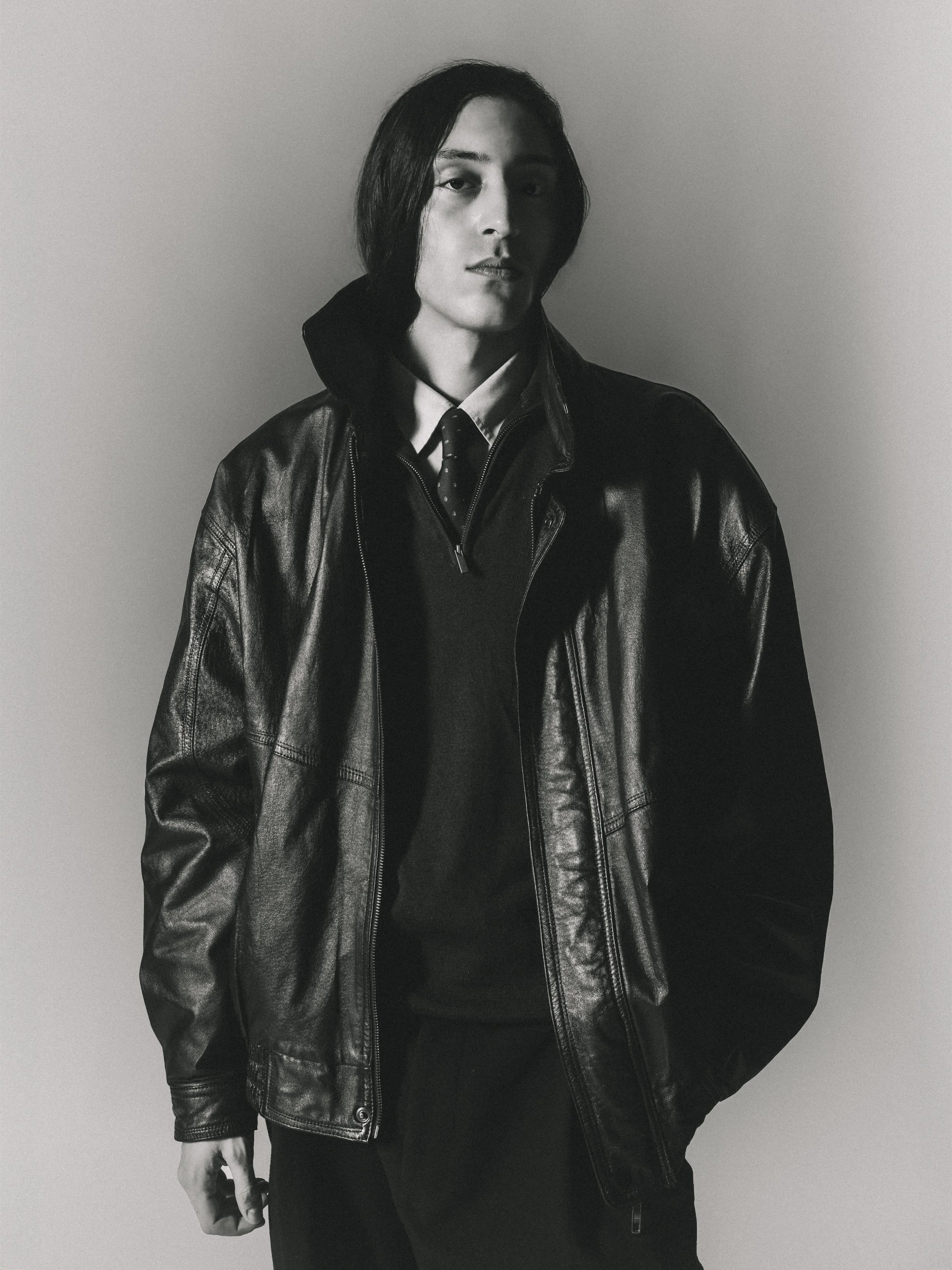 Une personne portant une veste en cuir noir, une chemise, une cravate et un pull noir, posant contre un mur gris. En noir et blanc.