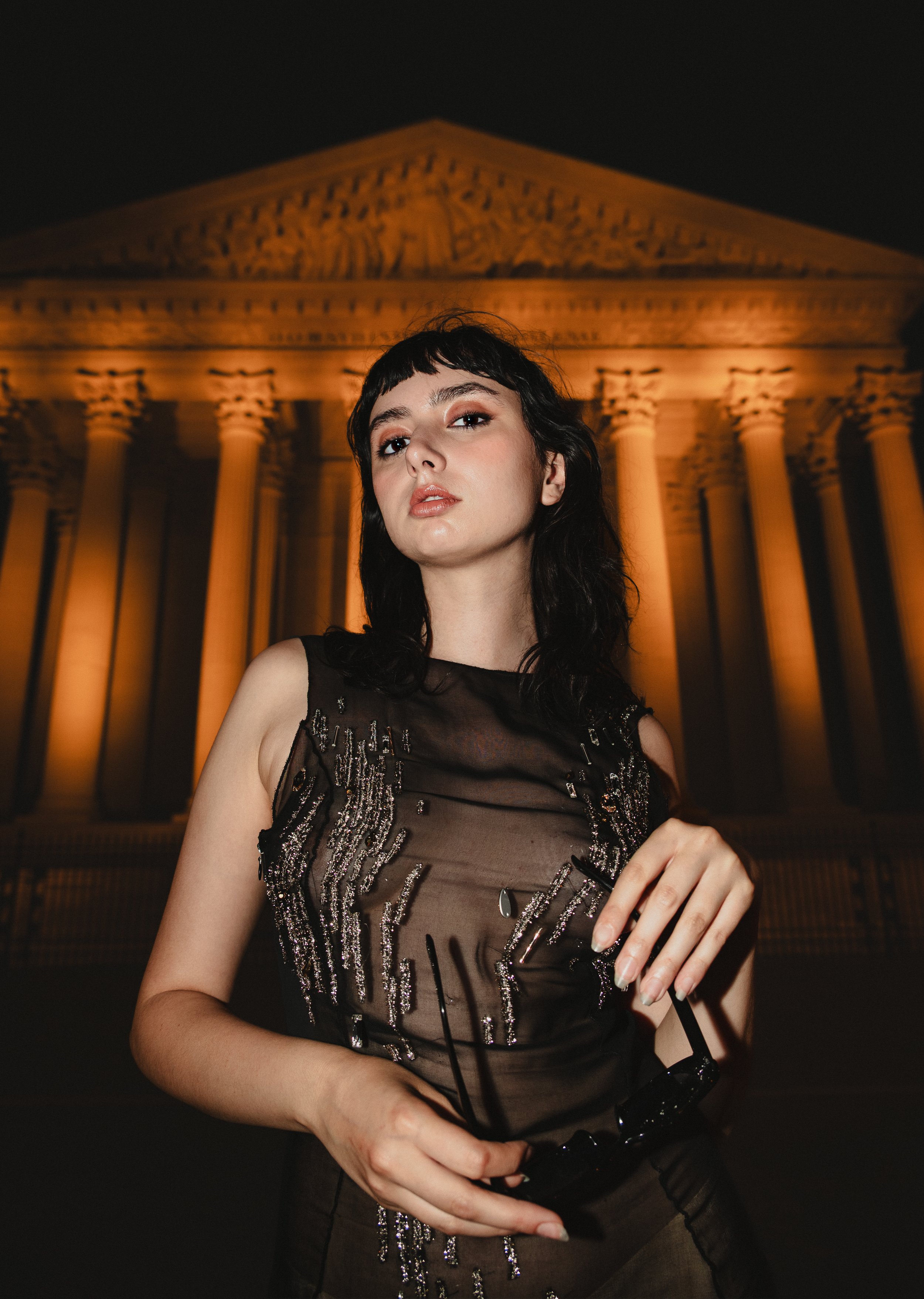 Une femme avec des cheveux noirs et une robe noire élégante se tient devant un bâtiment historique illuminé, ressemblant au Parthénon, en soirée.